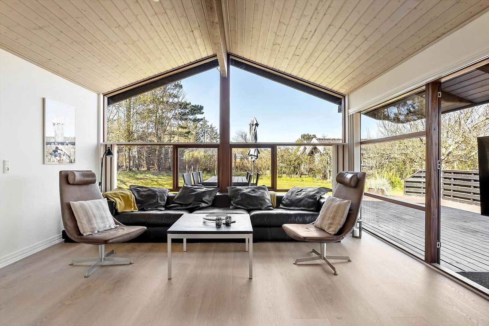 Wohnzimmer mit großem Fenster, Sofa und zwei Sesseln. Holzboden und hölzerner Deckenverkleidung.