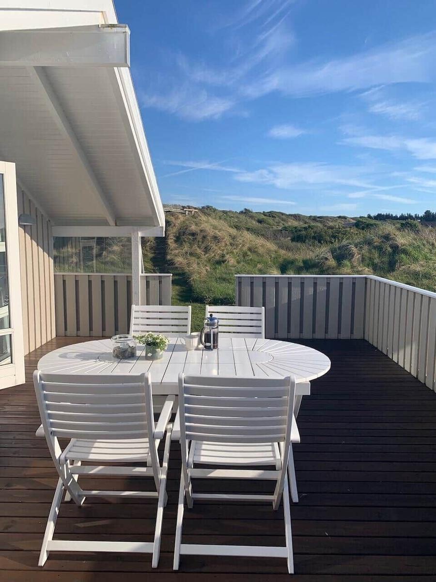 Terrasse mit weißem Tisch und Stühlen, Blick auf Dünen und blauen Himmel.
