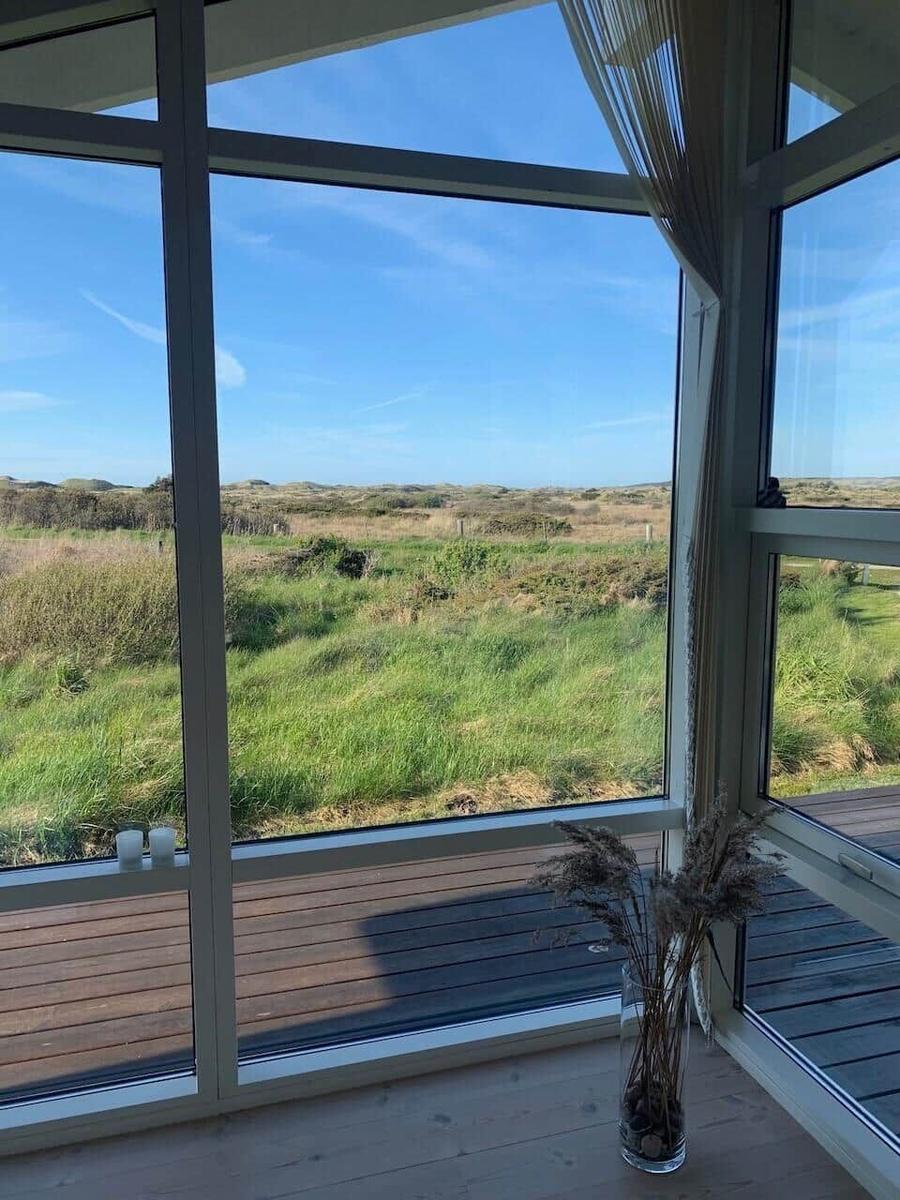 Großes Fenster mit Blick auf Grasland und blauen Himmel. Holzterrasse und Vasen mit Trockenpflanzen.