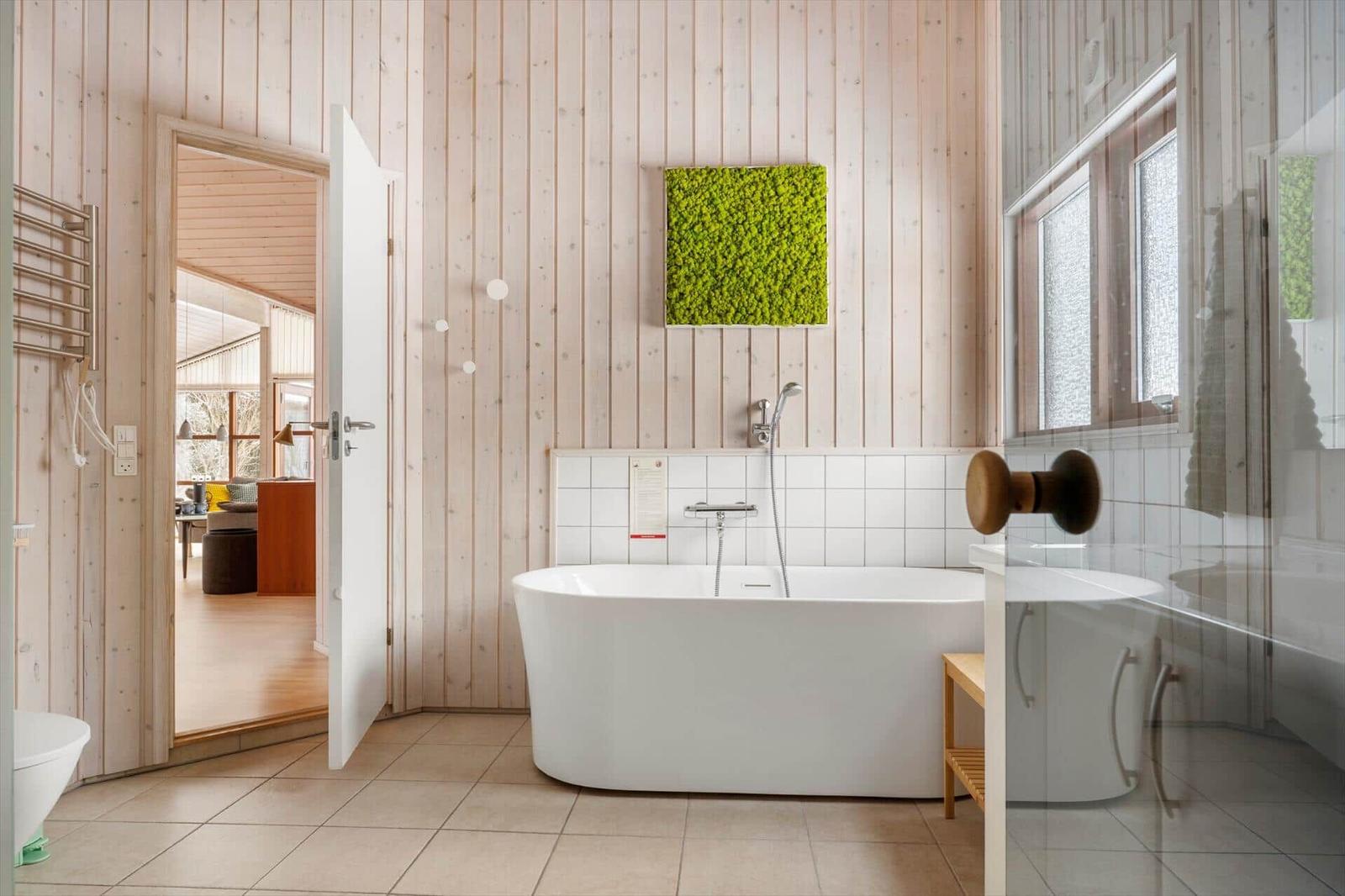 Badezimmer mit freistehender Badewanne, Holzwand und grünem Wandbild. Tür führt in Wohnbereich.