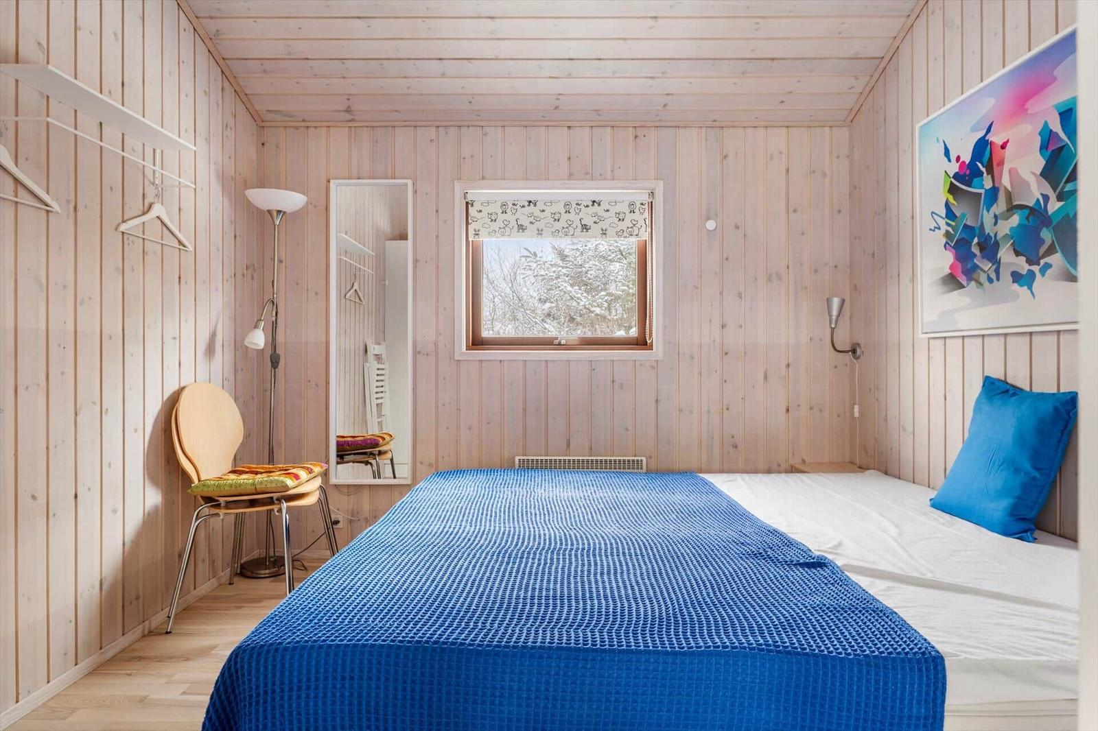 Schlafzimmer mit Holzwand, Bett mit blauer Bettdecke und Fenster mit Schneelandschaft.