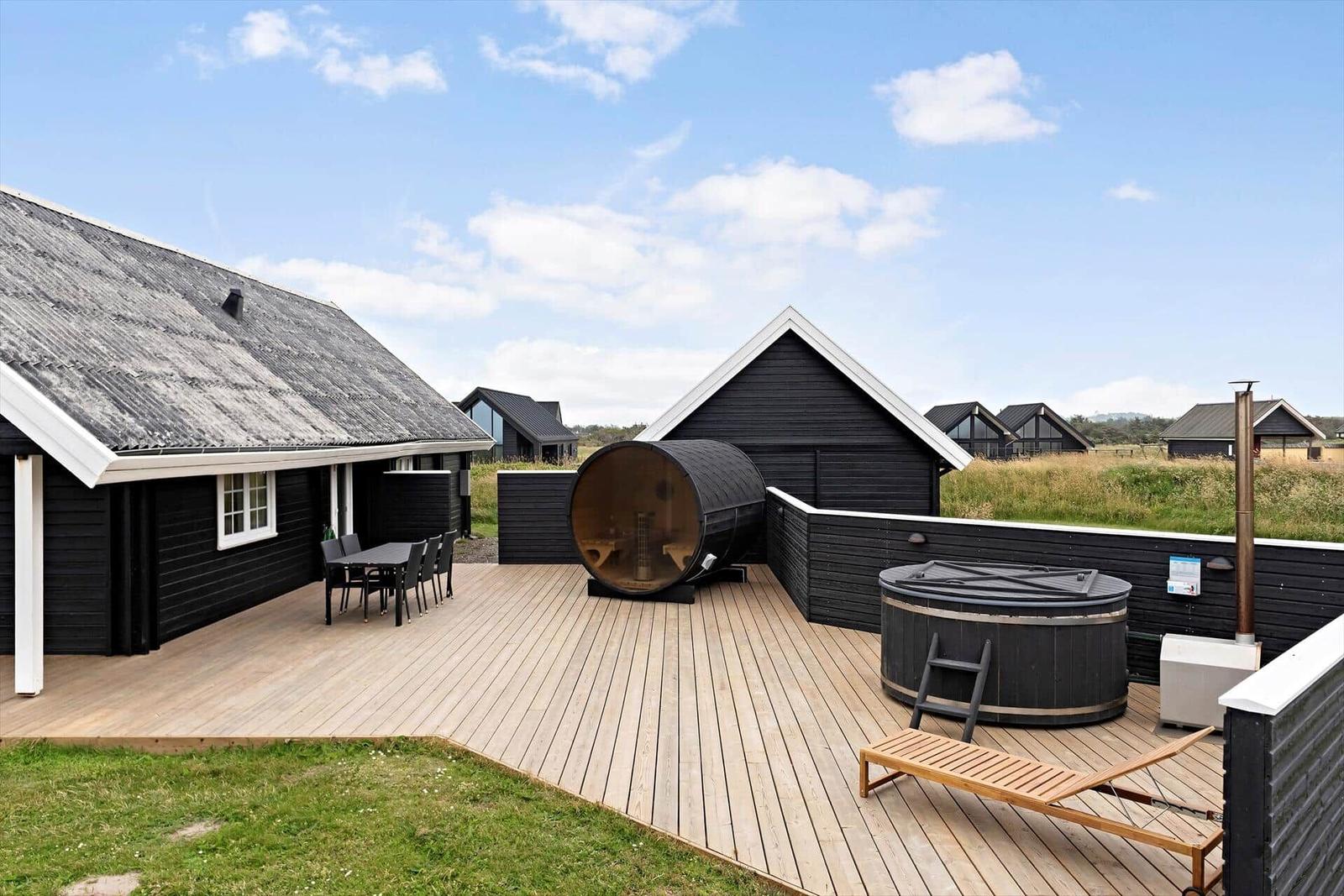 Terrasse mit Sauna, Whirlpool und Liegestuhl vor schwarzem Holzhaus.