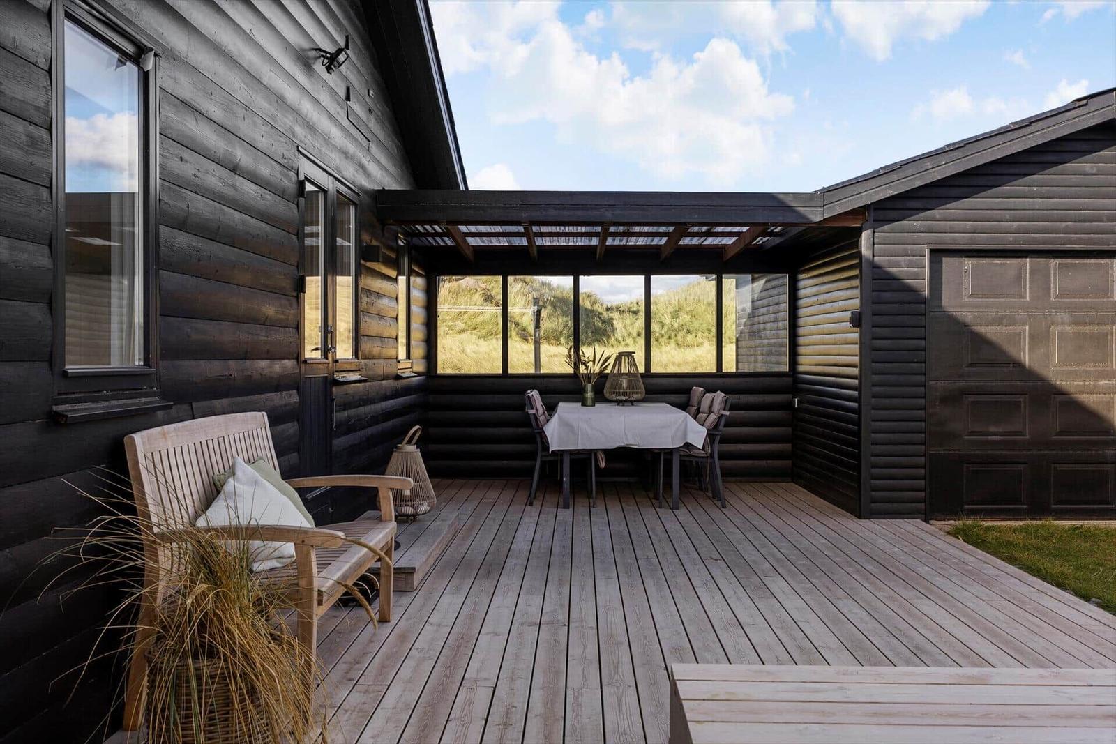 Terrasse mit Tisch, Stühlen und Sitzbank vor einem Holzhaus mit großem Fenster.