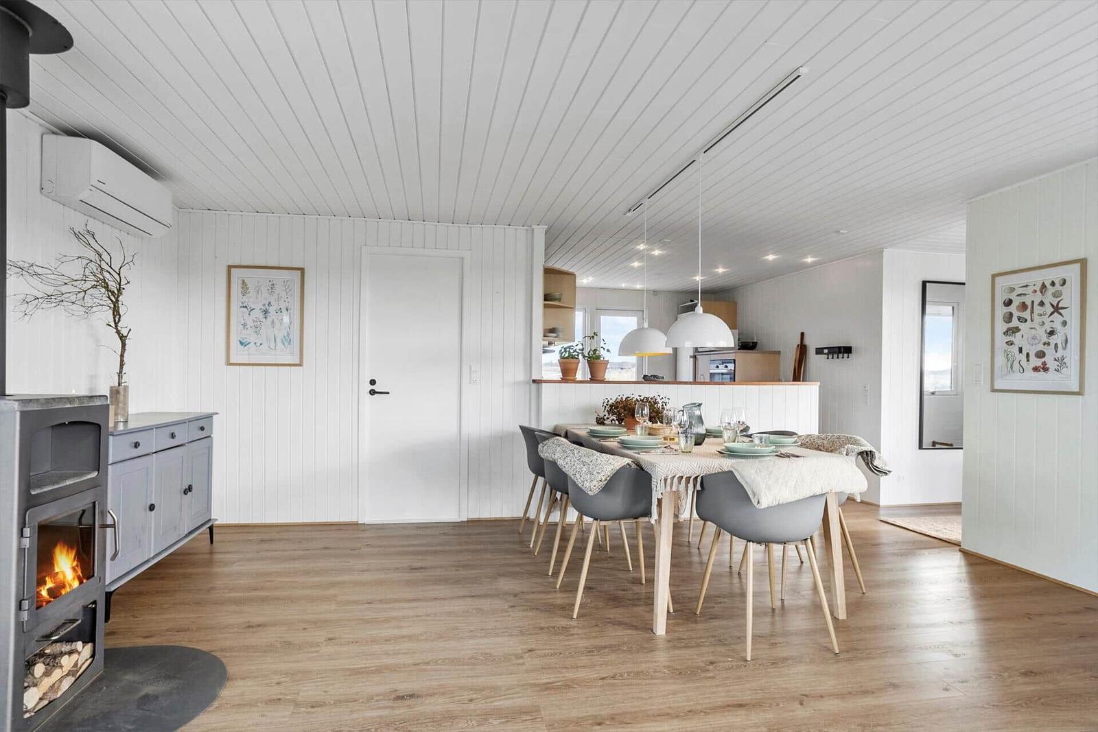 Essbereich mit Tisch, Stühlen und Kamin. Küche im Hintergrund. Weiße Wände und Holzboden.