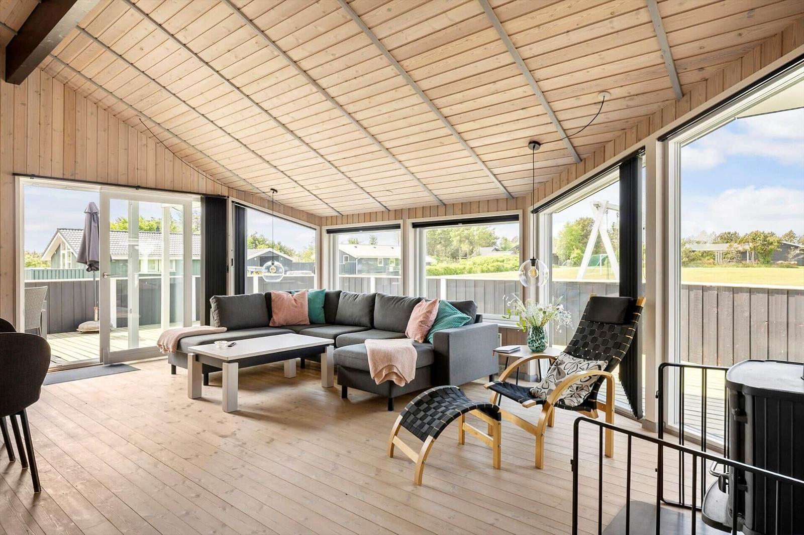 Ein Wohnbereich mit Holzboden, großem Sofa und vielen Fenstern. Draußen ist eine Terrasse mit Blick auf Grünflächen.