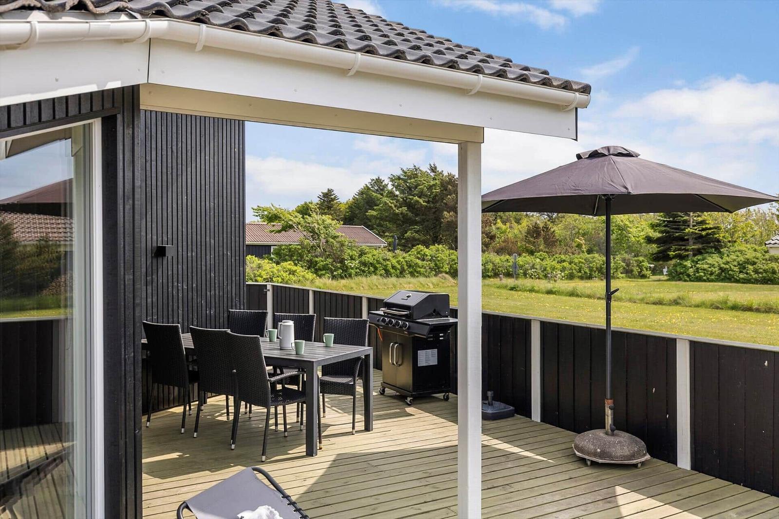 Terrasse mit Grill, Tisch, Stühlen und Sonnenschirm. Blick auf Grünfläche und Bäume.