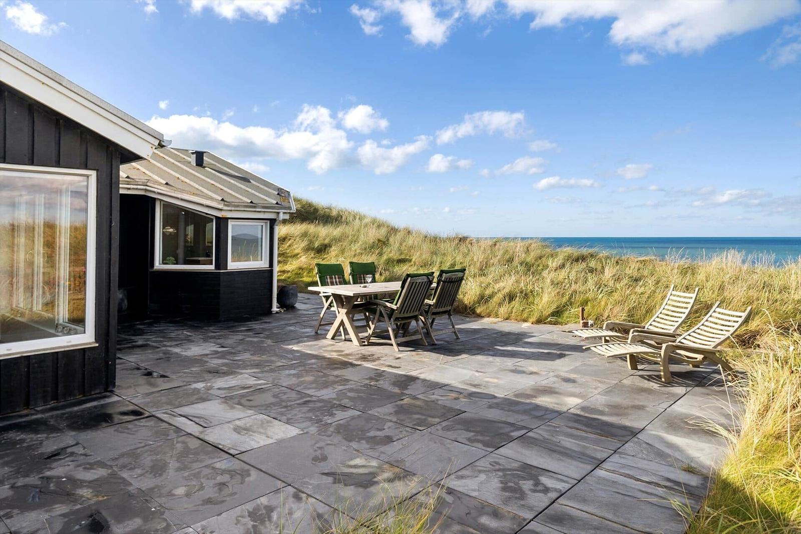 Terrasse mit Tisch, Stühlen und Liegestühlen. Blick auf das Meer über Dünen.