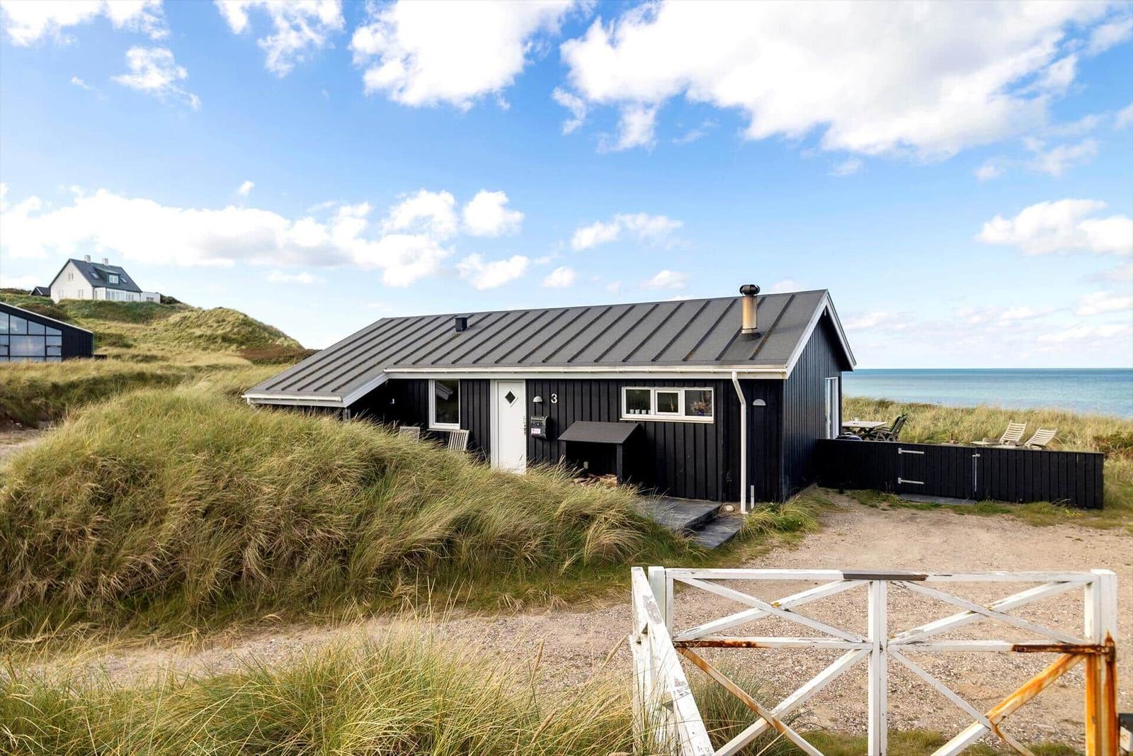 Schwarzes Strandhaus mit Terrasse, umgeben von Dünen und Meer.