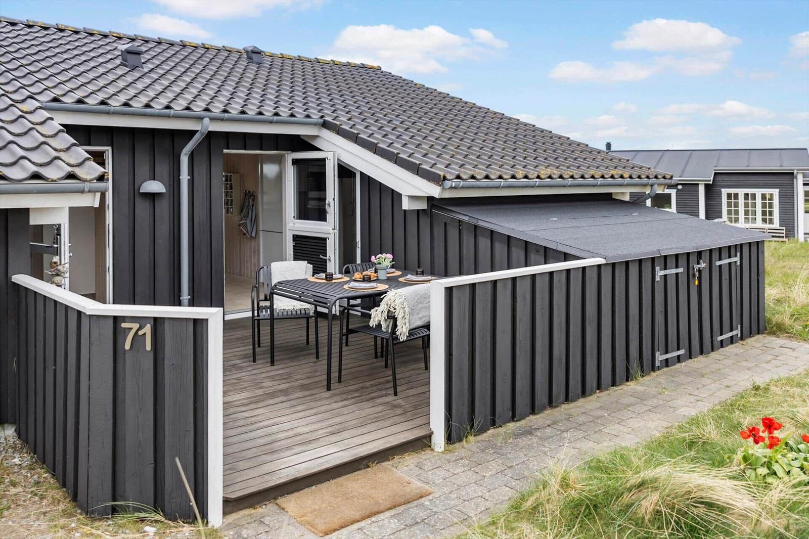 Haus mit Terrasse und Tisch. Schwarze Fassade, Holzdecke, Hausnummer 71.