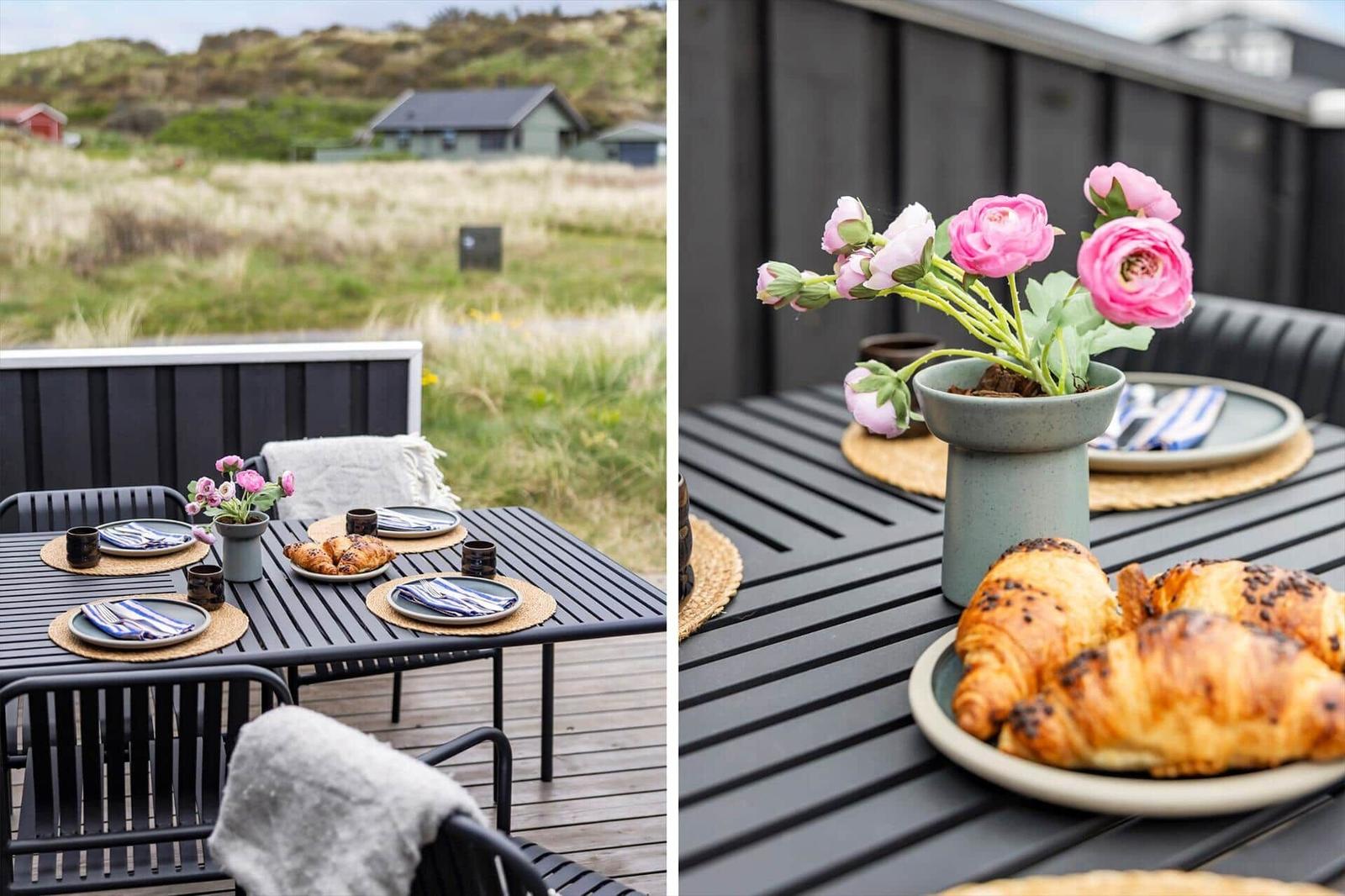 Terrasse mit Tisch, Stühlen und Frühstück. Hintergrund: Grasland und Häuser.