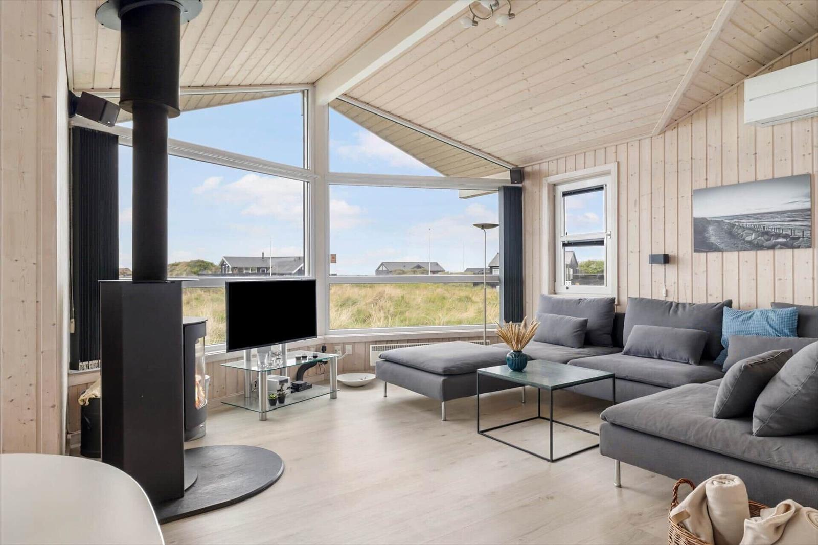 Wohnzimmer mit Holzwänden, großem Fenster und grauem Sofa. Holzdecke und Kaminofen sichtbar.