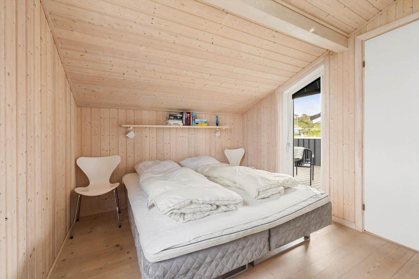 Schlafzimmer mit Holzwand, Bett und Stuhl. Fensterblick auf Terrasse.