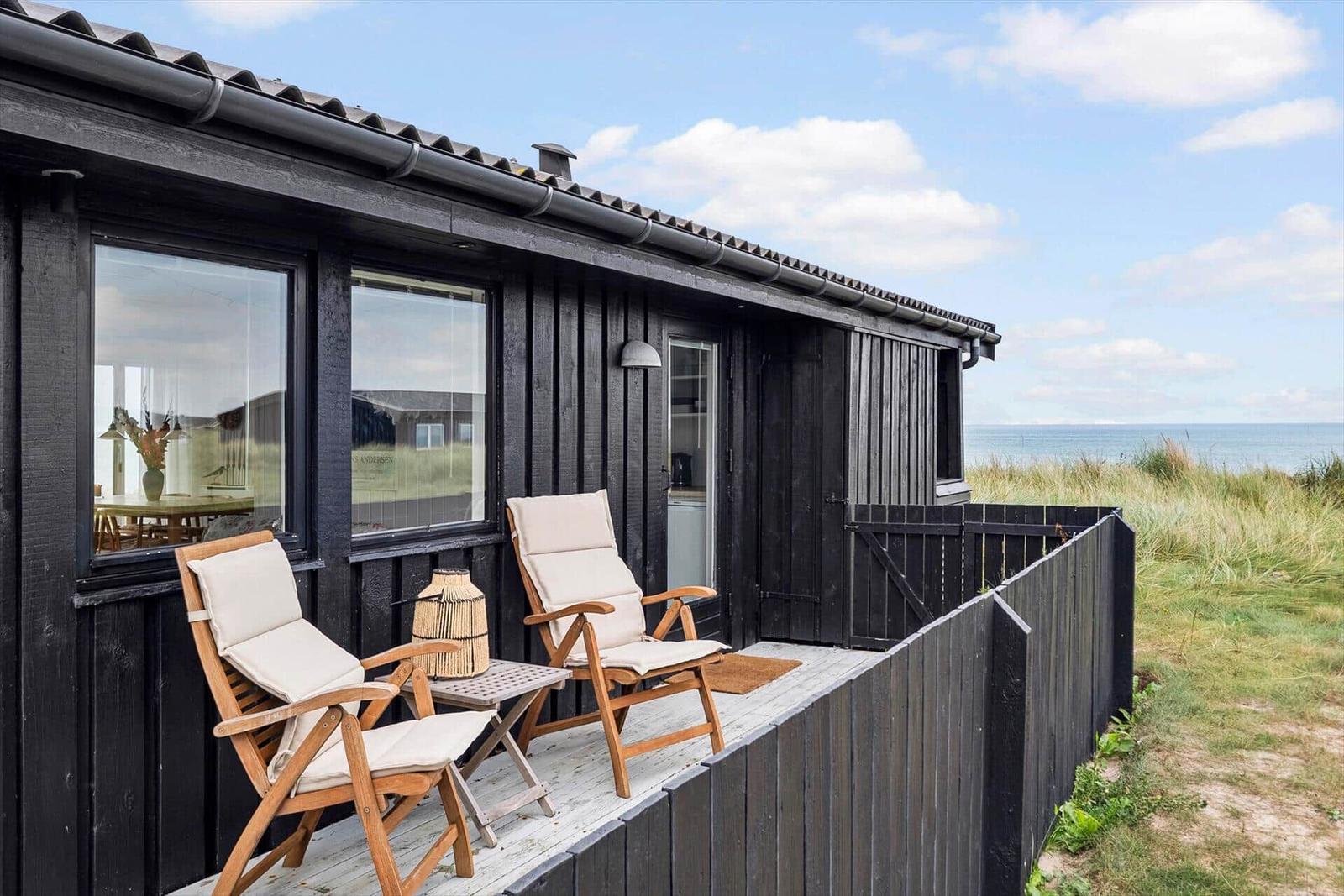 Schwarzes Strandhaus mit Terrasse, zwei Holzstühlen und Meerblick.