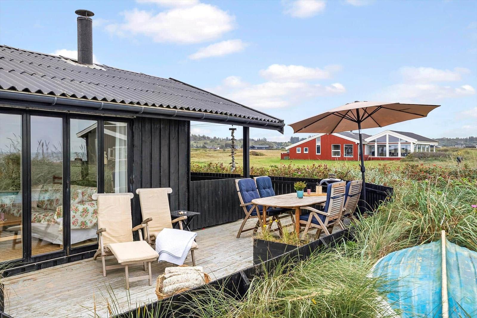 Terrasse mit Sitzgelegenheiten und Sonnenschirm. Im Hintergrund rotes Haus und Grasland.