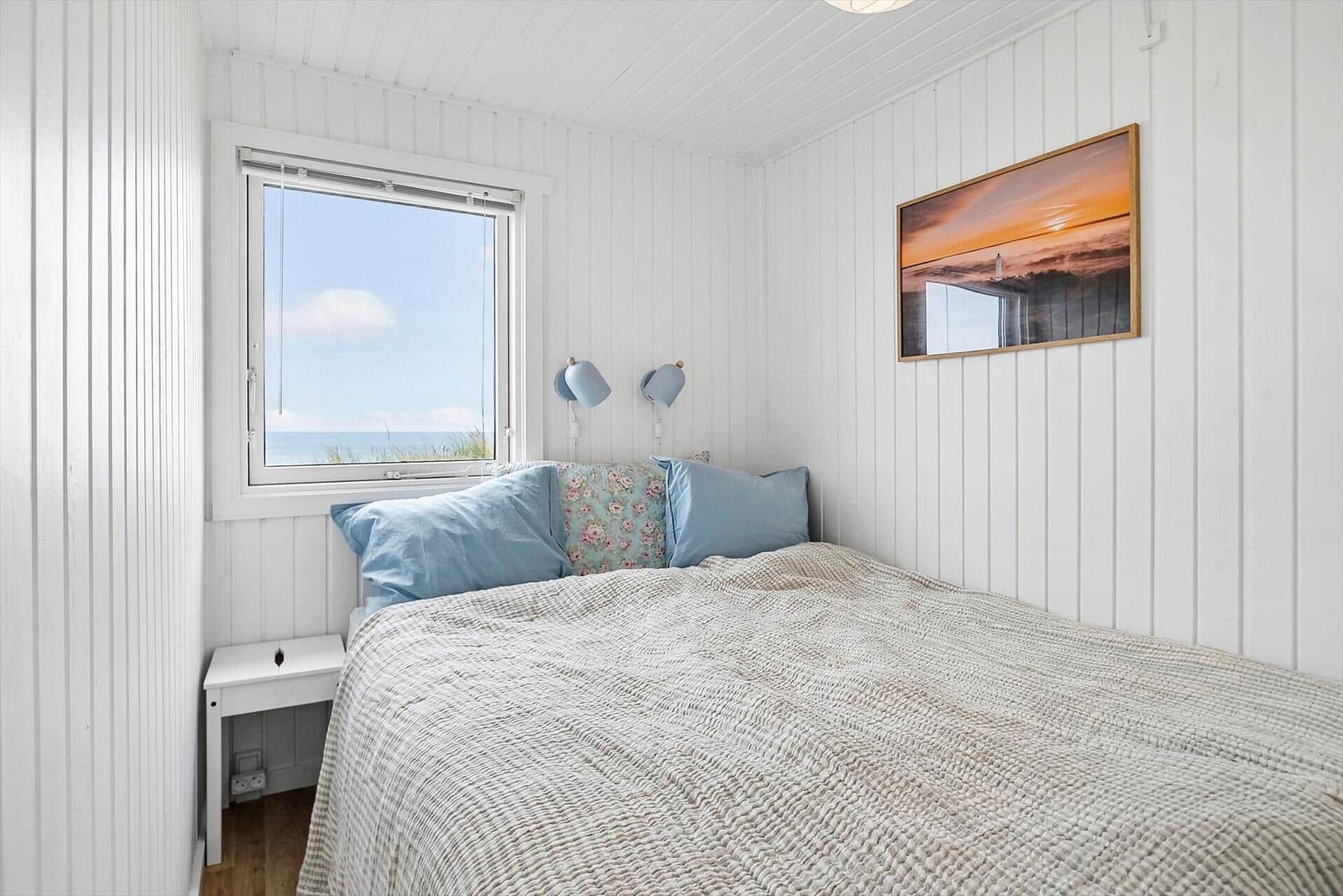 Schlafzimmer mit Blick aufs Meer, weiß verkleidete Wände, Bett mit Kissen und Wandbild.
