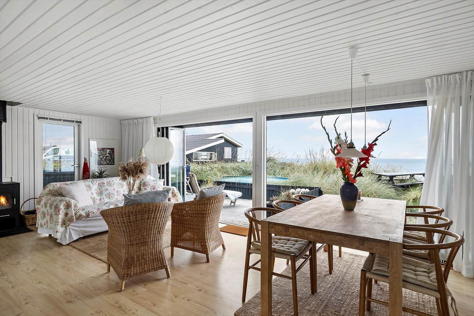 Wohn- und Essbereich mit Blick aufs Meer und Garten. Holztisch, Stühle, Sofa und Kamin.