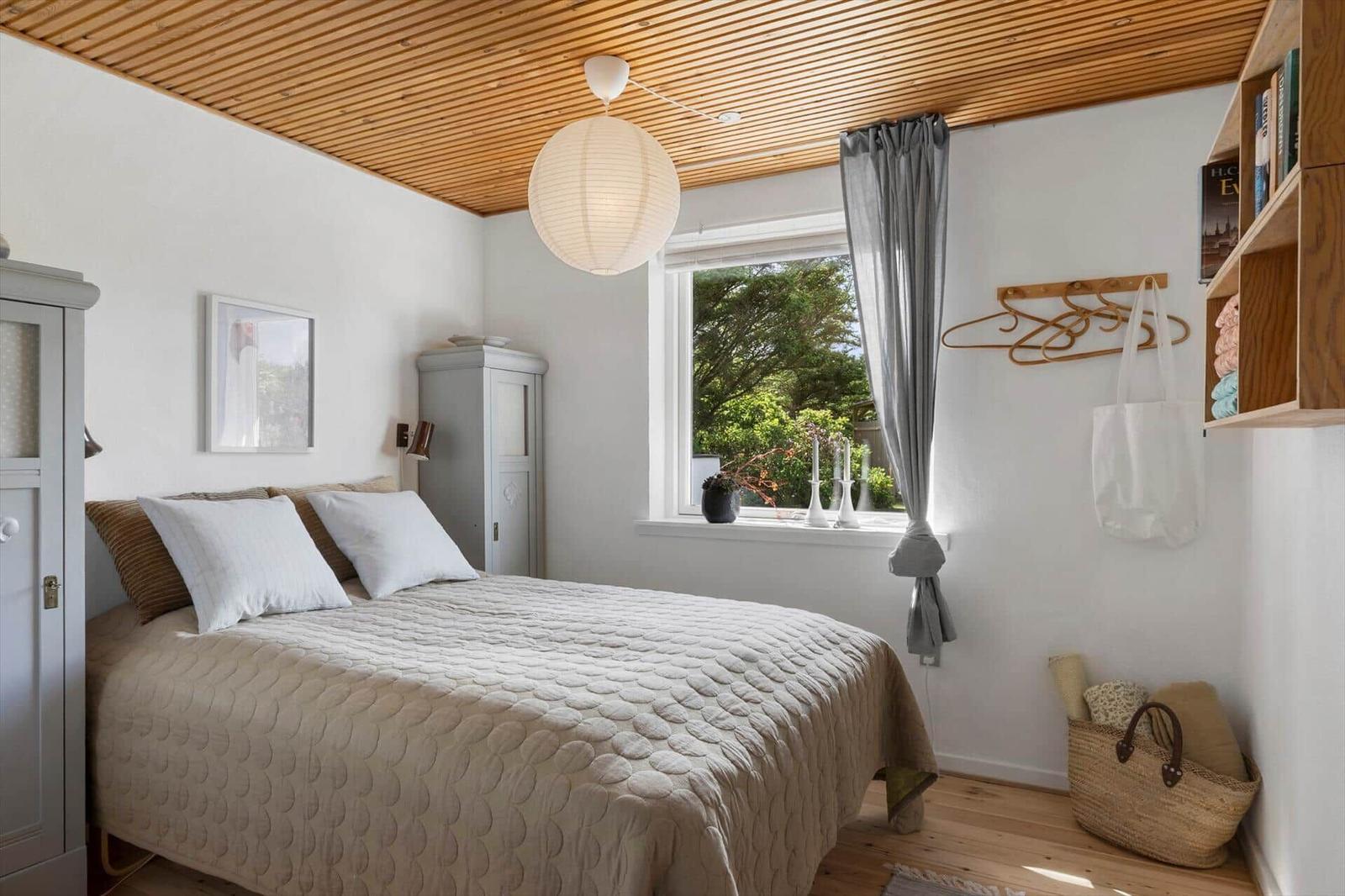 Schlafzimmer mit Bett, Fenster, Holzdecke und Schrank. Wandregal und Lampe sind sichtbar.