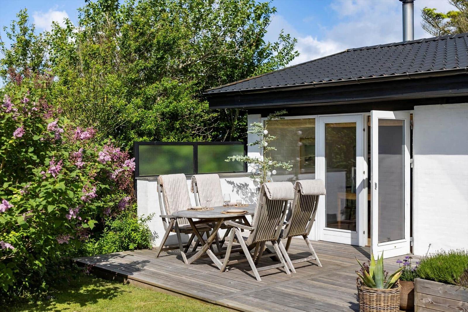 Terrasse mit Holztisch und Stühlen, umgeben von Grün und Blumen.