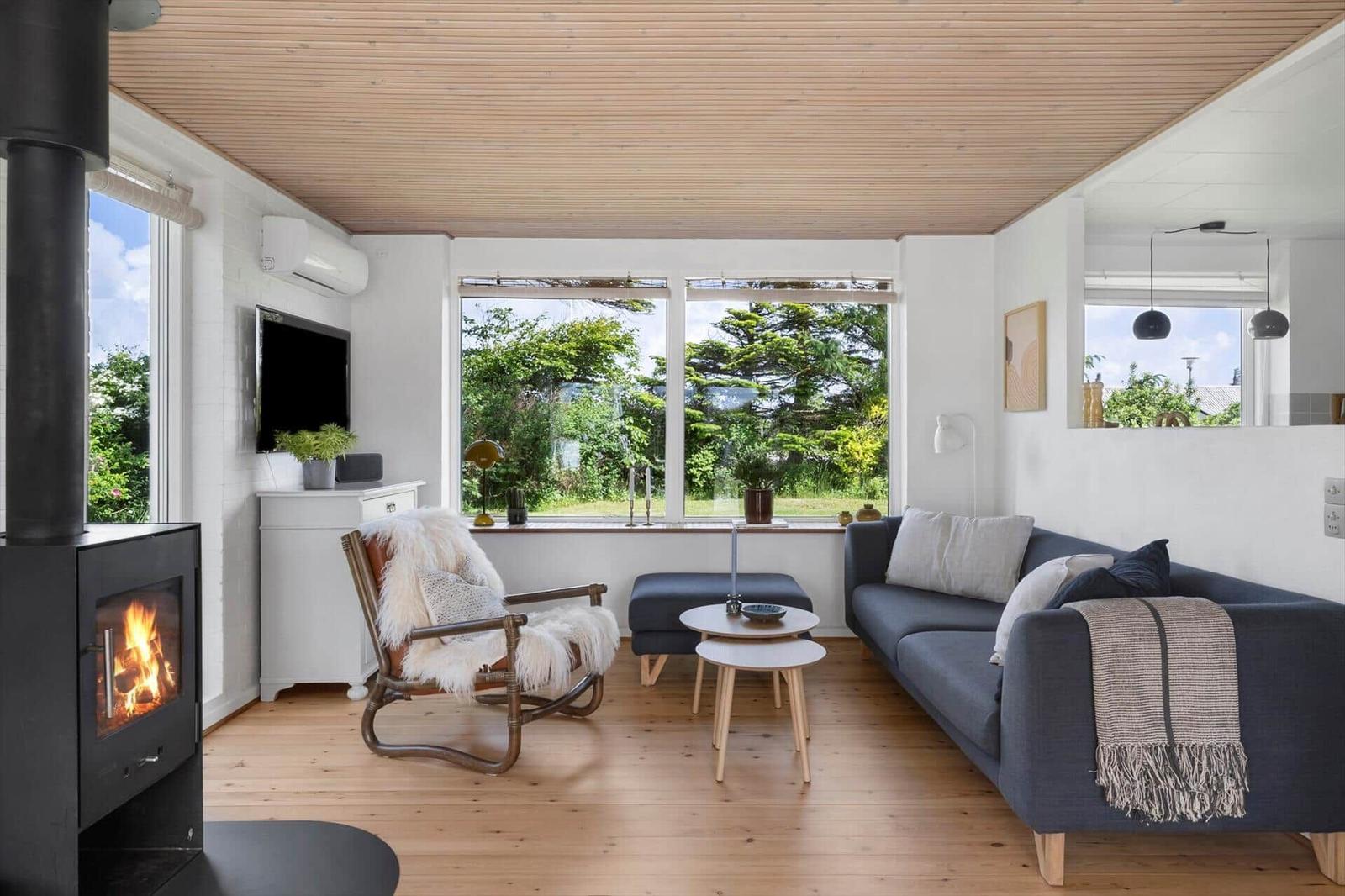 Wohnzimmer mit Kamin, Sofa und Sessel. Große Fenster bieten Blick auf Garten.