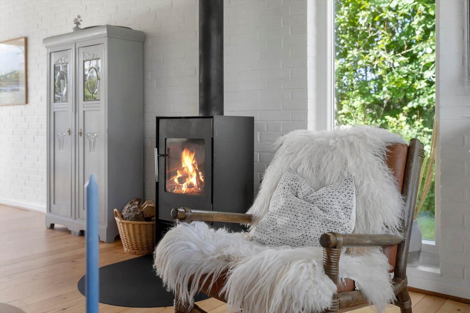 Ein Kaminofen mit brennendem Holz steht neben einem Stuhl mit Felldecke und Kissen.