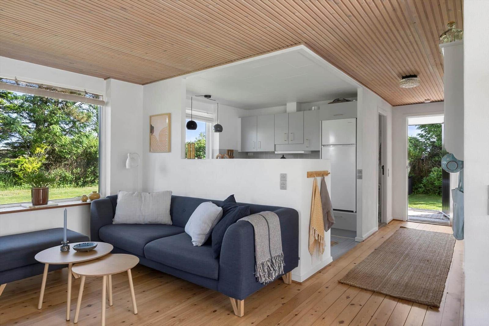Wohnzimmer mit Sofa, Tischen und Blick auf Garten. Küche mit Kühlschrank sichtbar.