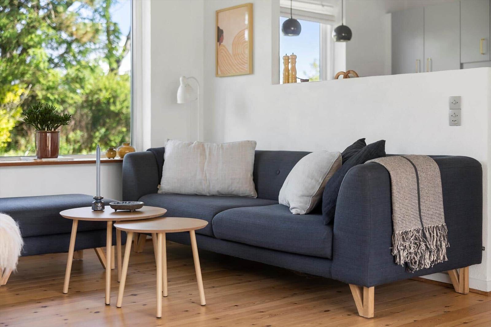 Ein Wohnzimmer mit grauem Sofa, zwei Holztischen und Blick auf Bäume.