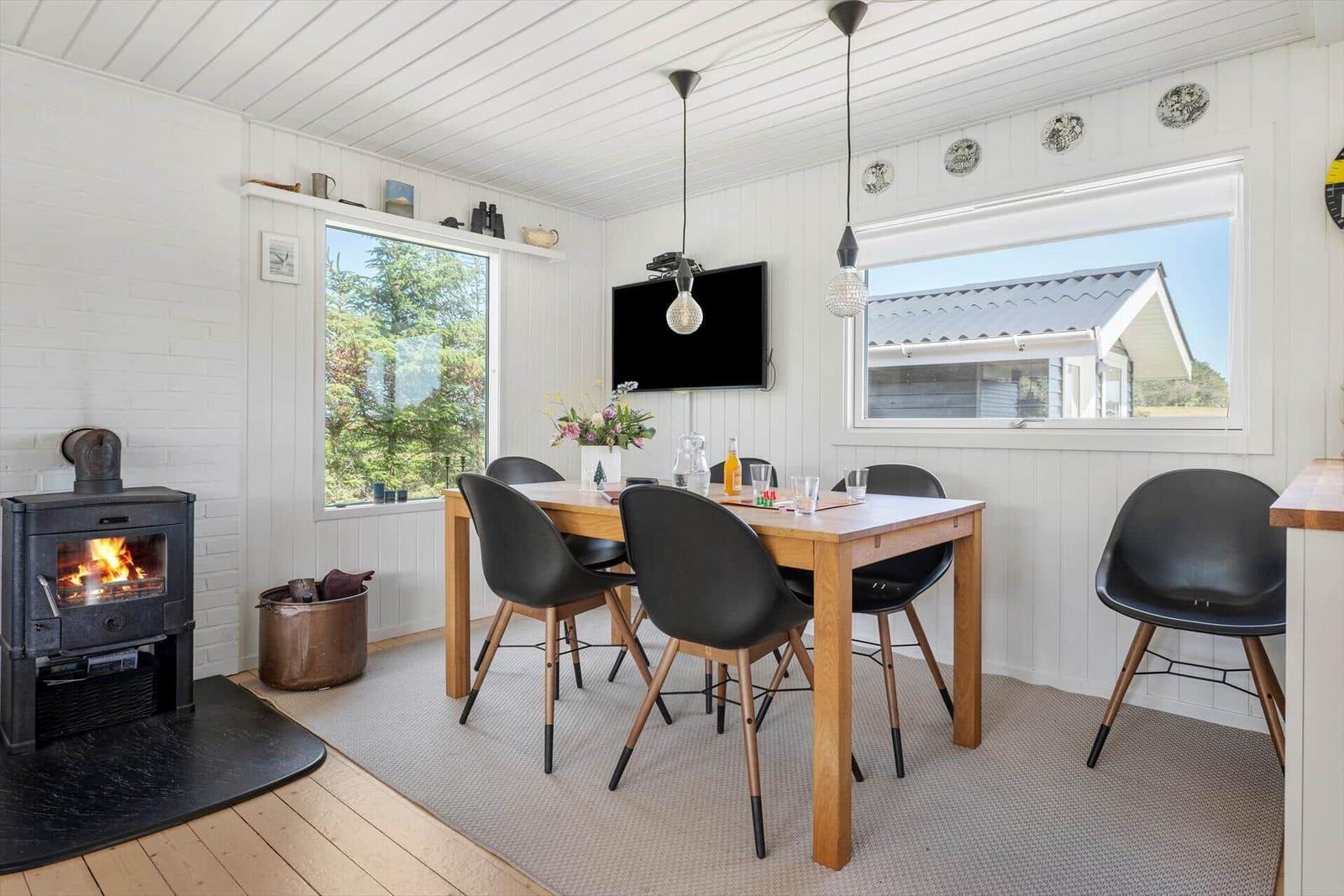 Esszimmer mit Holztisch, schwarzen Stühlen und Kaminofen. Fenster zeigen Wald und Haus.