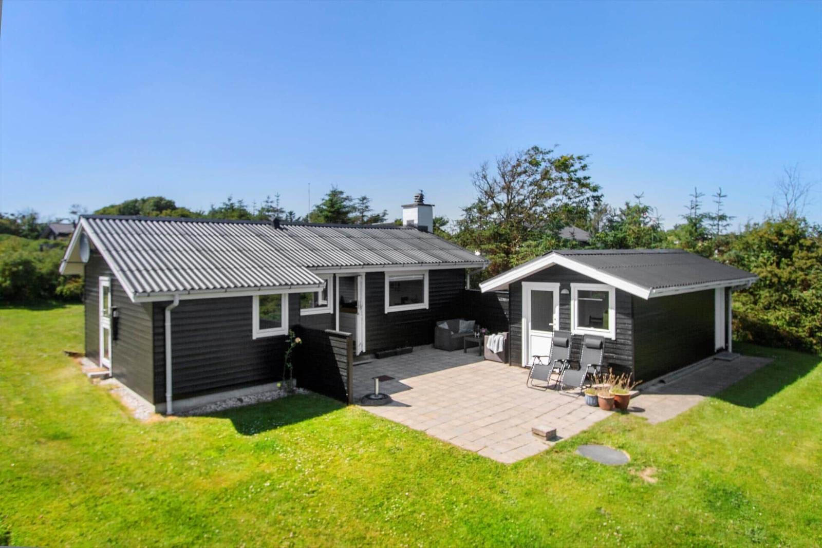 Schwarze Holzhütte mit Terrasse und Garten. Weiße Fenster und Türen. Hintergrund mit Bäumen.
