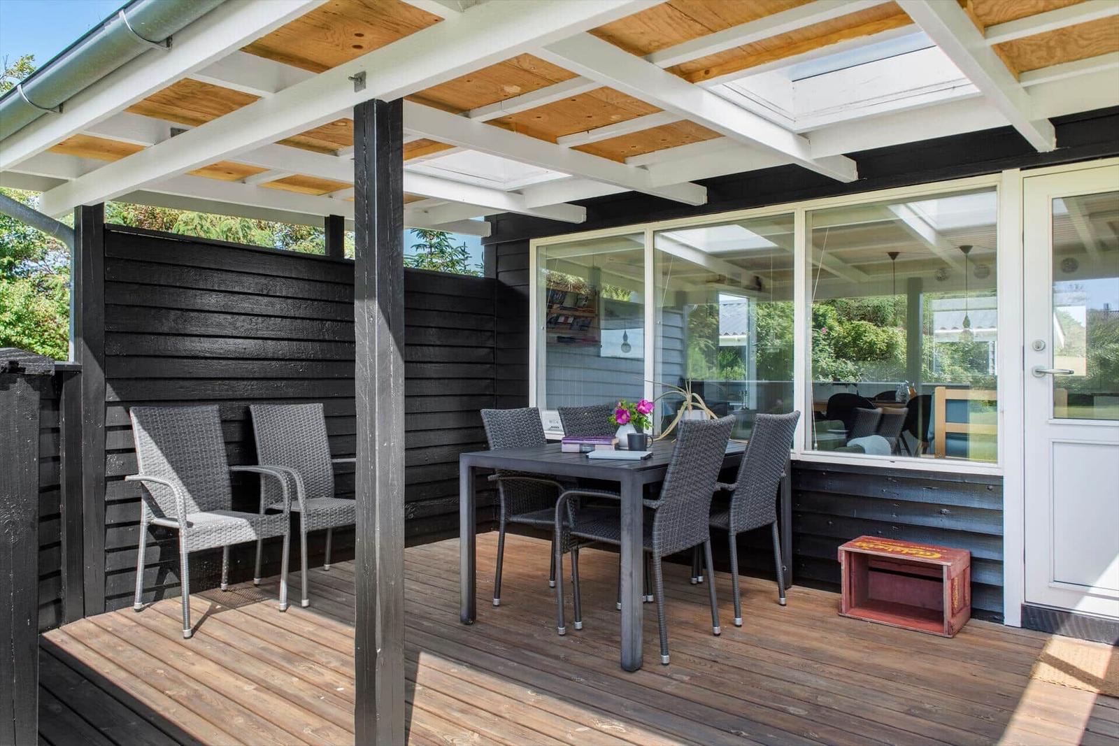 Terrasse mit Tisch, Stühlen und Dach aus Holz und Metall. Glasfront und roter Kistenkasten sichtbar.
