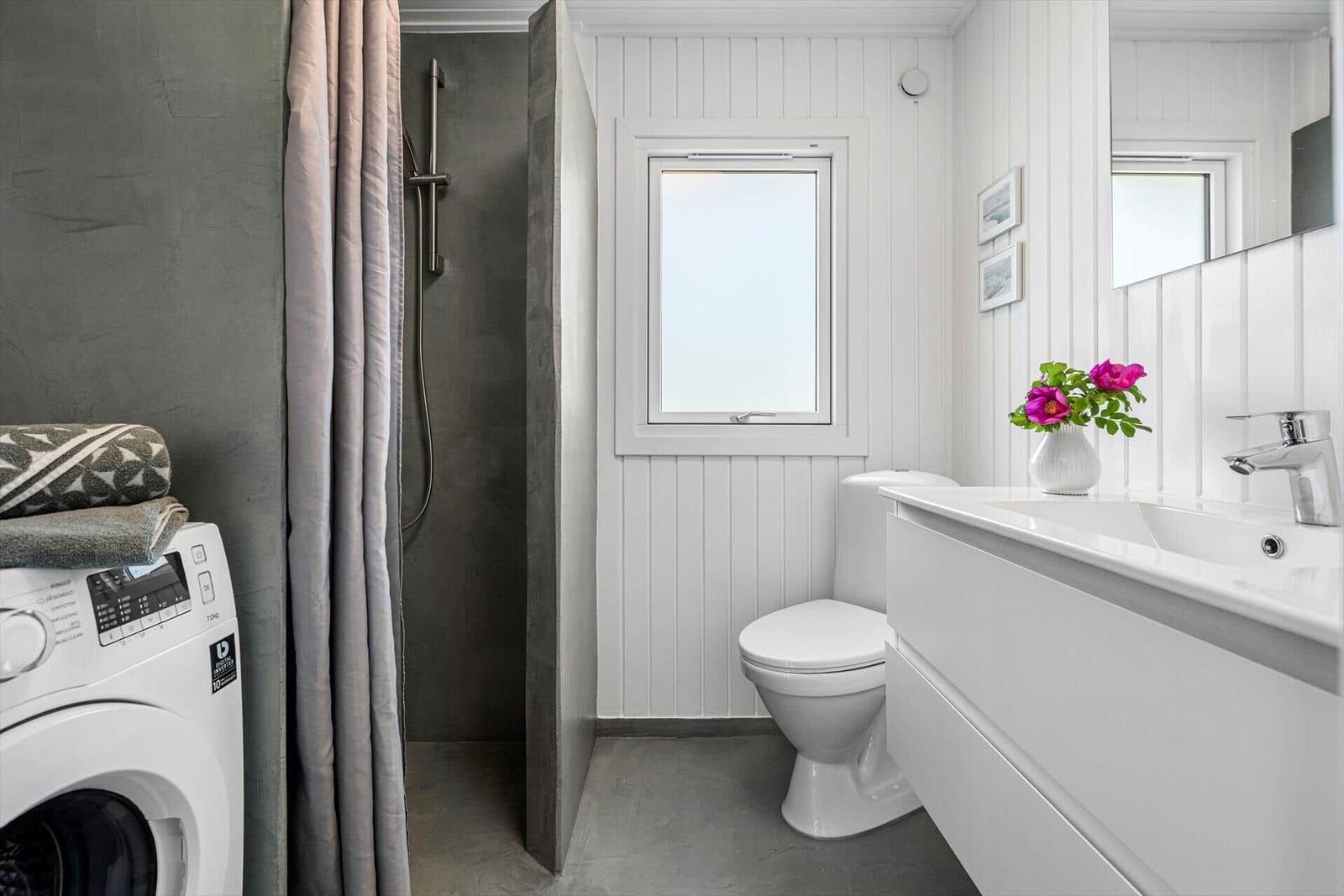 Badezimmer mit Dusche, Toilette und Waschbecken. Fenster, Spiegel und Blumen sind sichtbar.