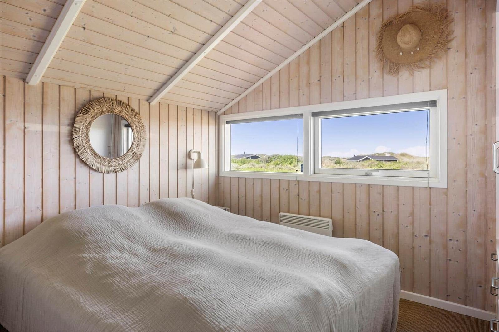Schlafzimmer mit Holzwand und Dach, Bett mit weißer Bettwäsche, Fenster mit Blick auf Landschaft.