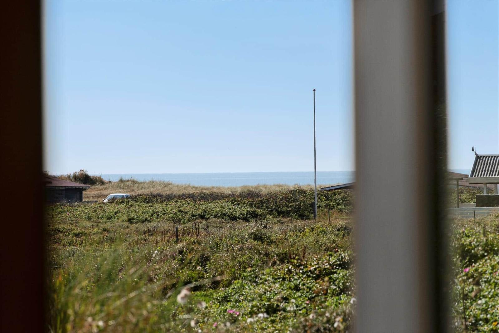 Durch ein Fenster blickt man auf eine Wiese mit Gras und Blumen, dahinter das Meer und einen Mast.