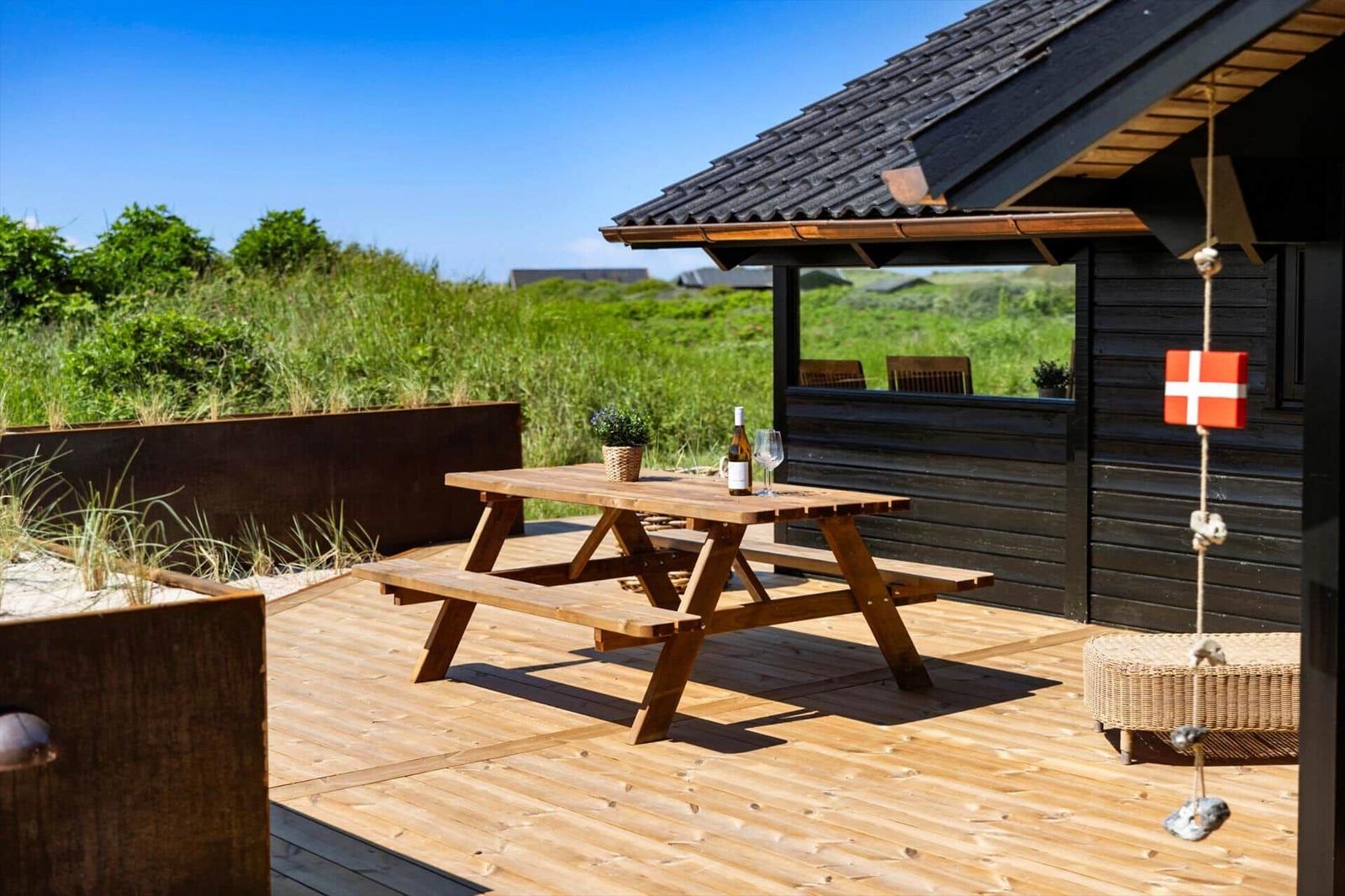 Holzterrasse mit Picknicktisch, Wein und Pflanze. Hintergrund: grüne Wiesen und dunkler Holzaufbau.