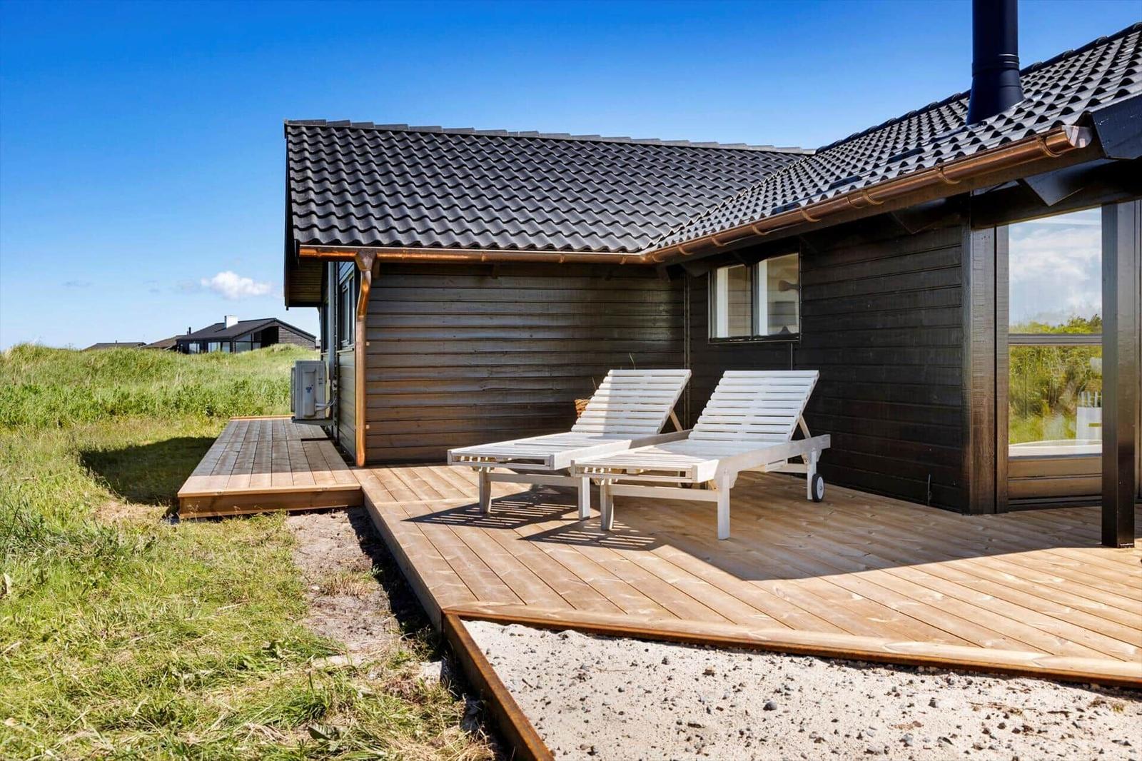 Holzterrasse mit zwei Liegestühlen vor einem dunklen Holzhaus mit Dach und Fenster.