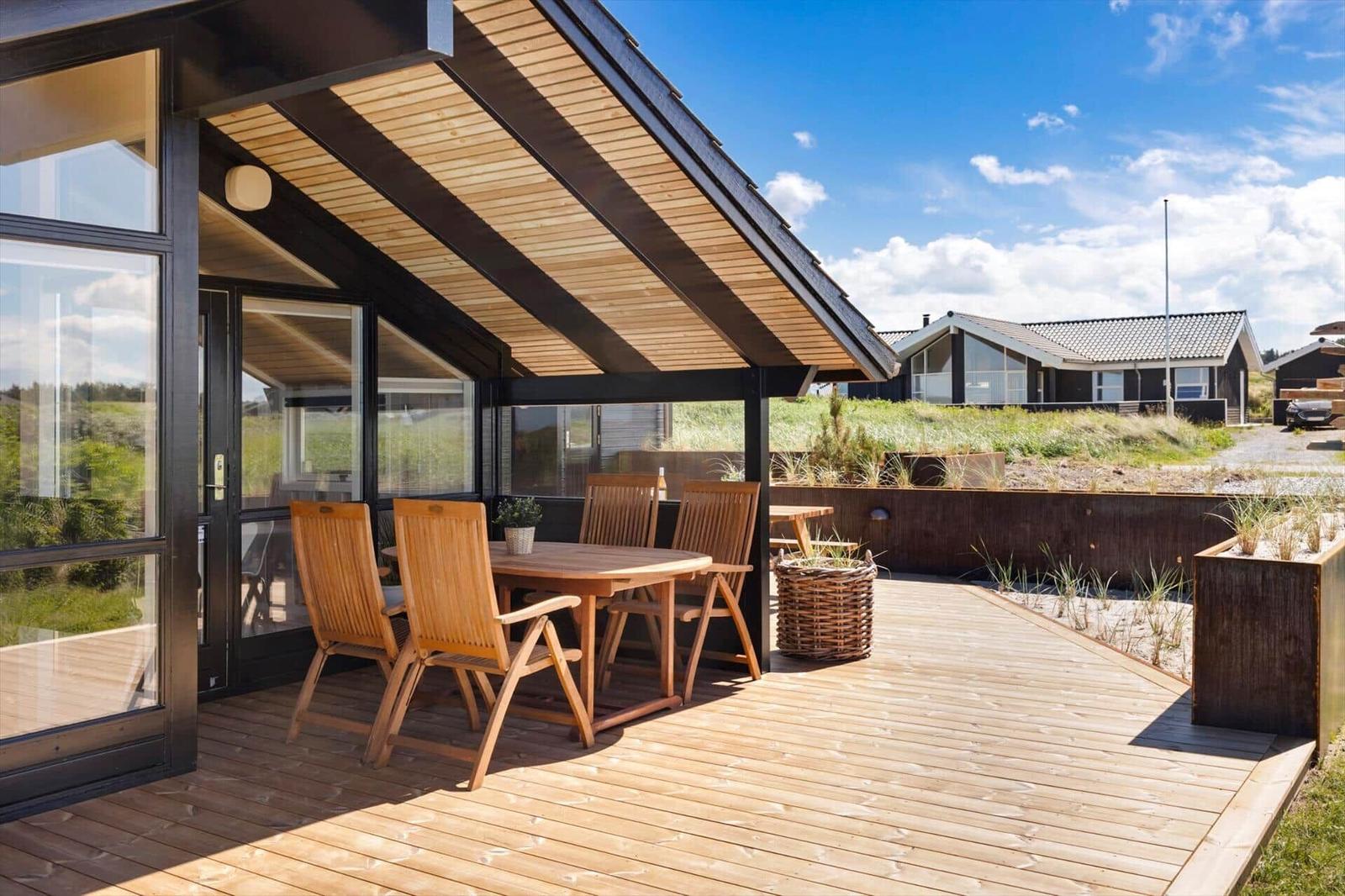 Terrasse mit Holztisch und Stühlen unter Dach. Holzdecke und Pflanzen im Hintergrund.