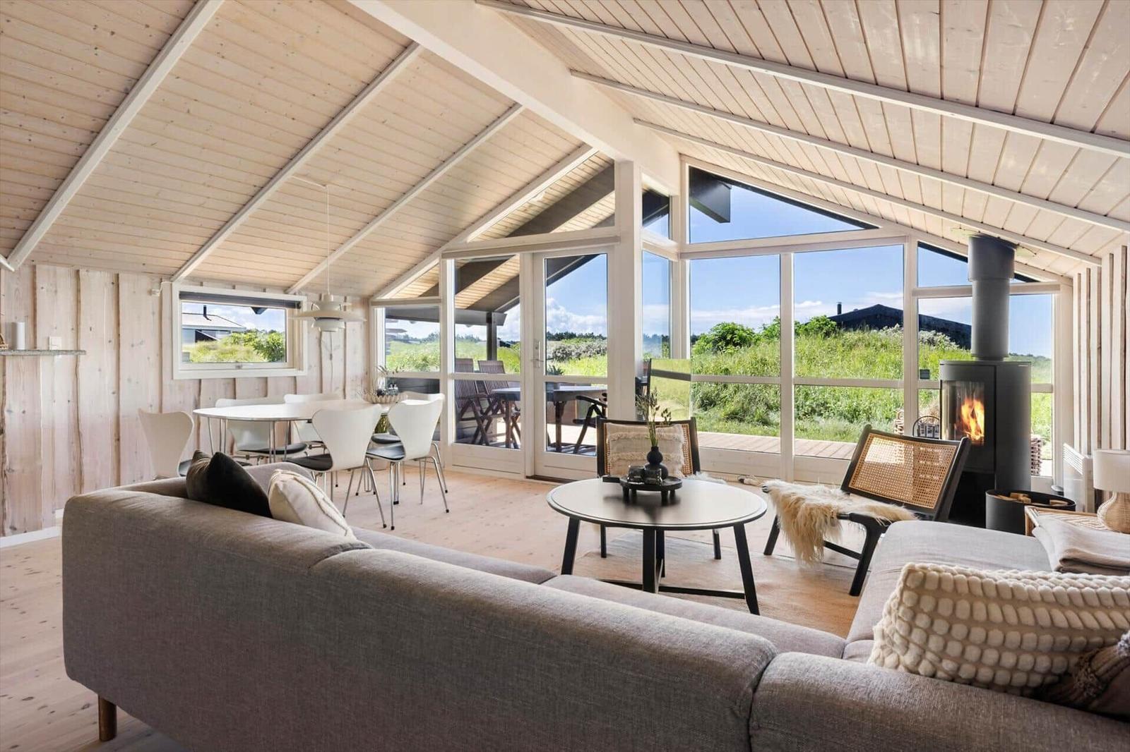 Ein Wohnzimmer mit Sofa, Esstisch und Kamin. Große Fenster bieten Blick auf die Landschaft.