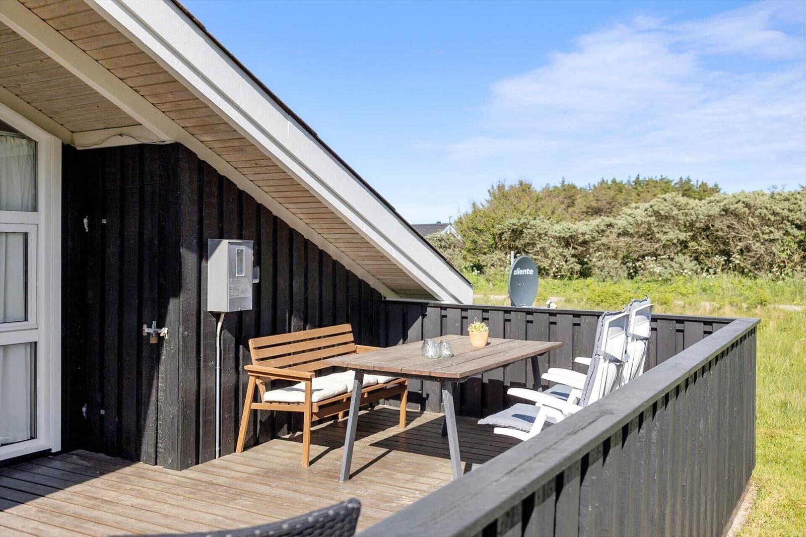 Terrasse mit Tisch, Bänken und Liegestühlen. Hintergrund: Grünflächen und Satellitenschüssel.