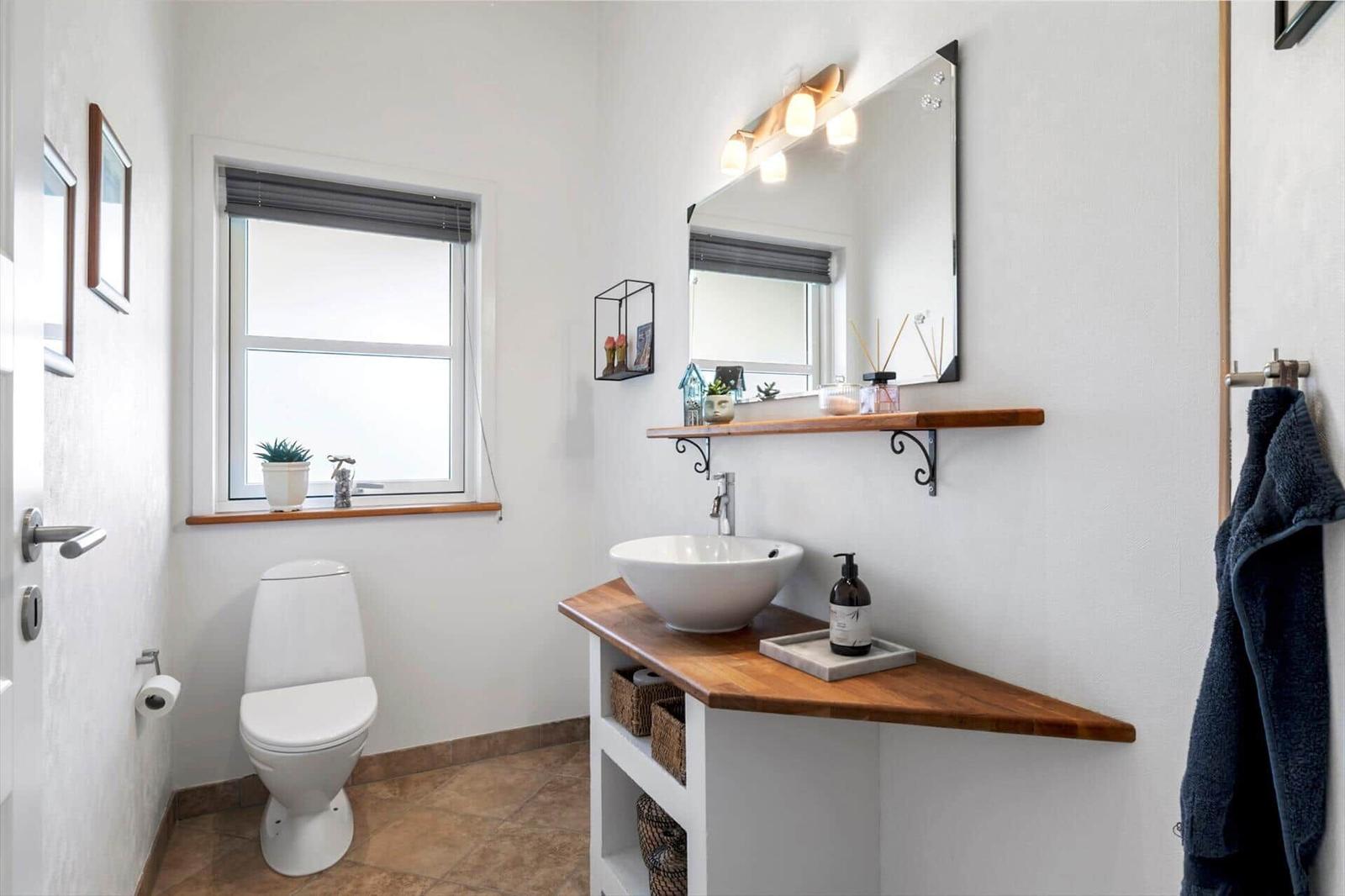 Badezimmer mit Waschbecken, Spiegel und Toilette. Holzregal und Fenster mit Pflanze.