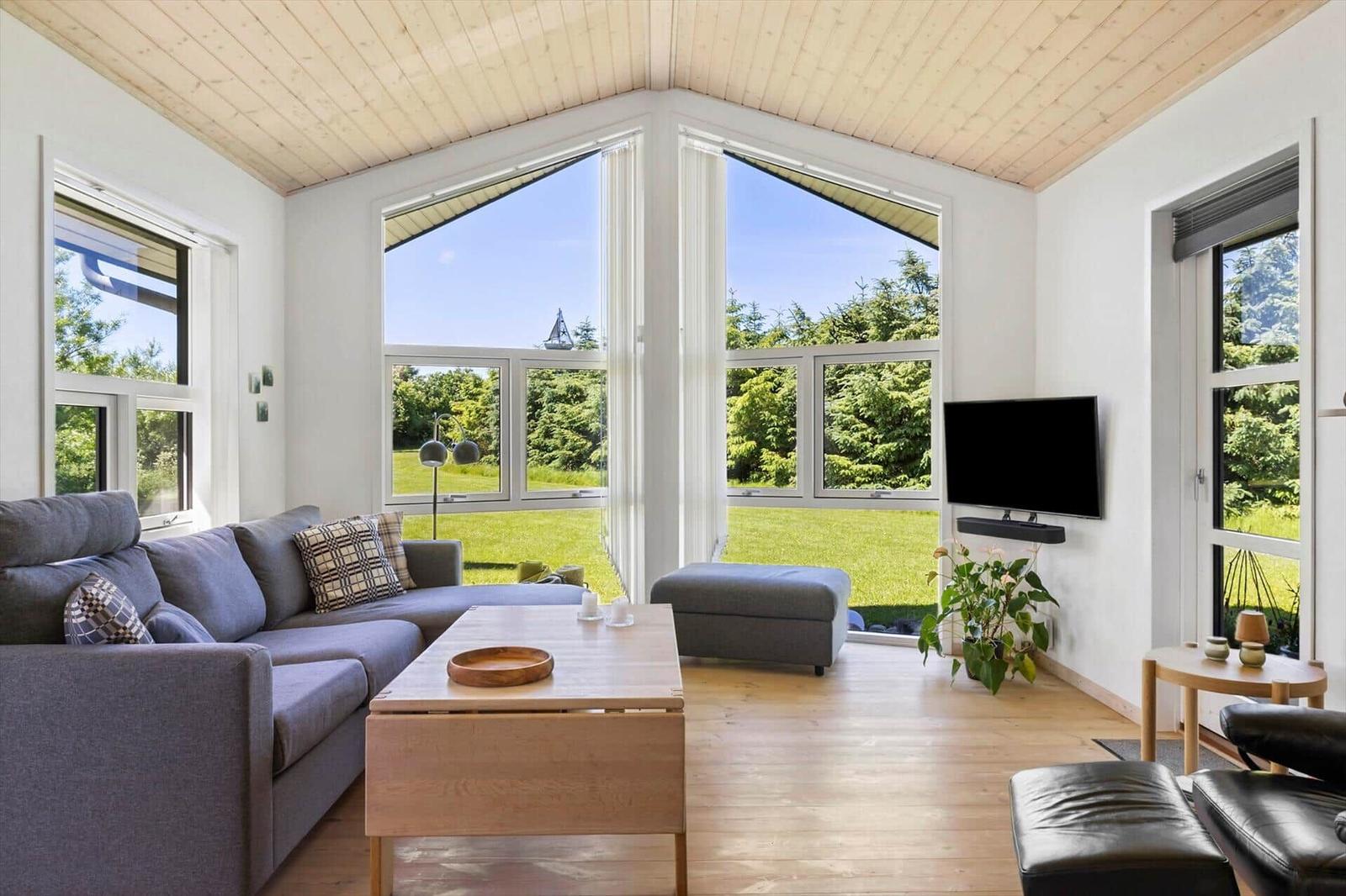 Wohnzimmer mit Sofa, Couchtisch und TV. Große Fenster bieten Blick auf Garten.