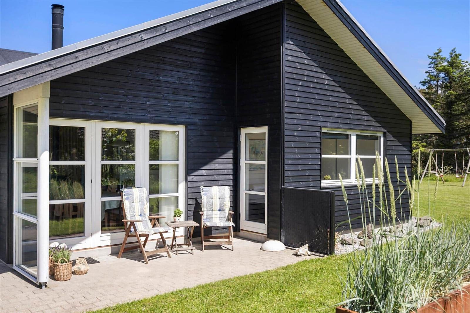 Schwarzes Holzhaus mit Terrasse, zwei Stühlen und Garten. Hinterhof mit Schaukel.