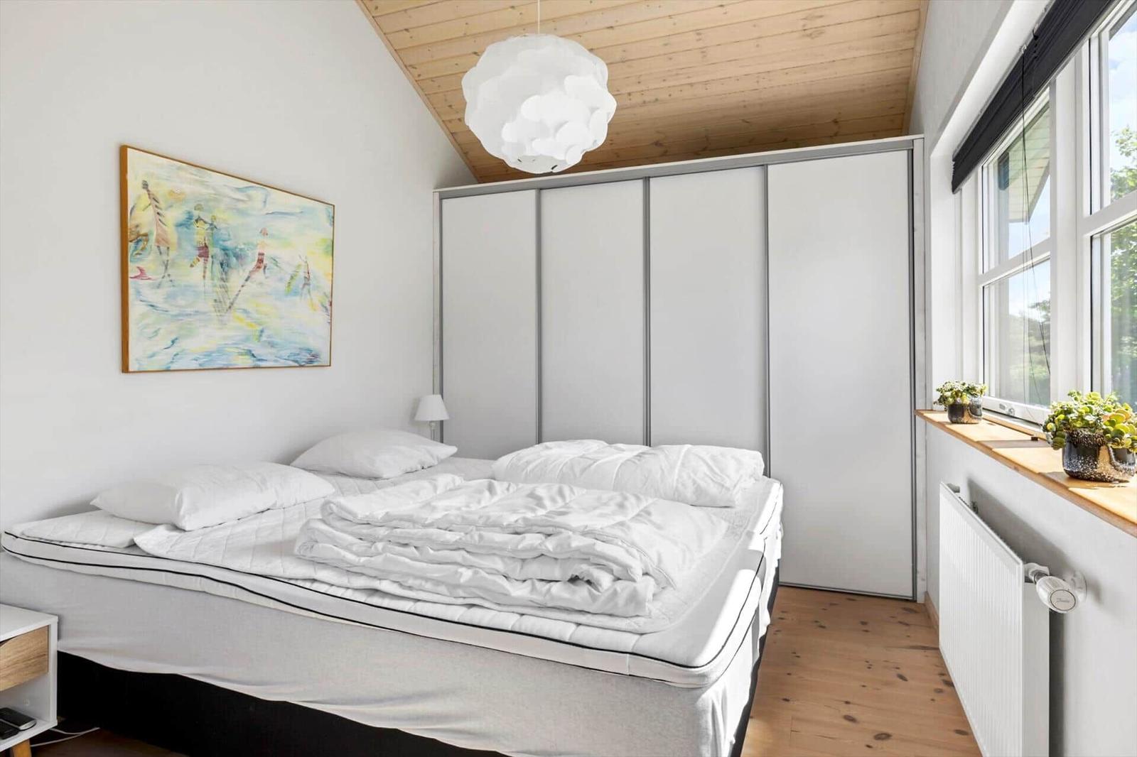 Schlafzimmer mit Bett, Schrank und Fenster. Decke aus Holz, Wandbild und Lampe.
