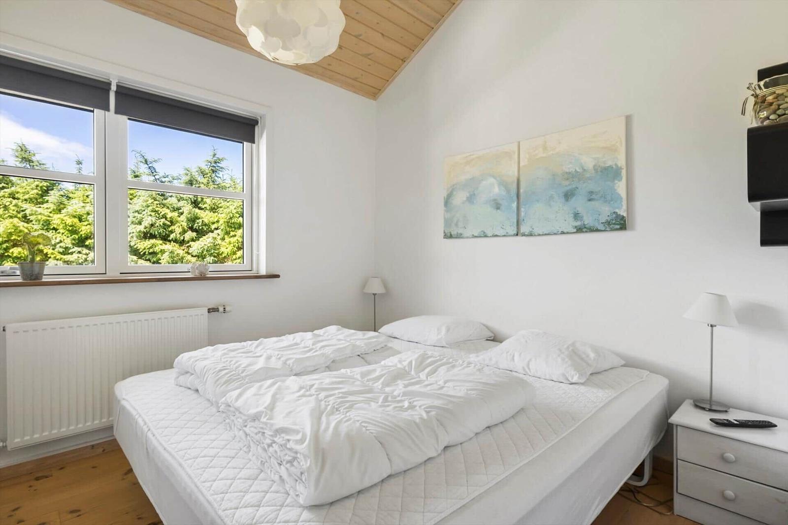 Schlafzimmer mit weißem Bett, Fenster mit Blick auf Bäume und Holzdecke.