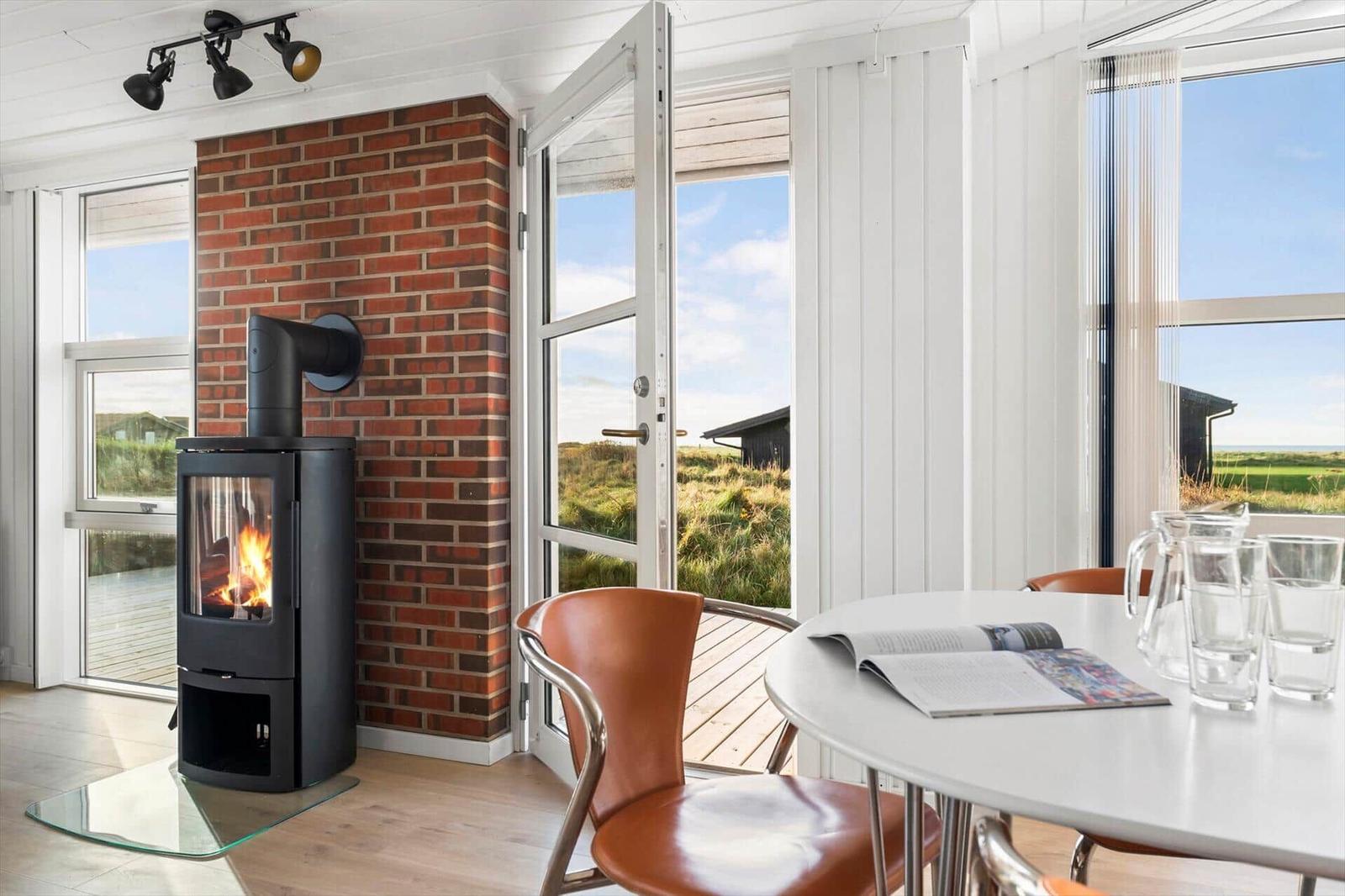 Ein Raum mit Kamin, Tisch und Stühlen. Durch die Fenster ist eine Landschaft zu sehen.