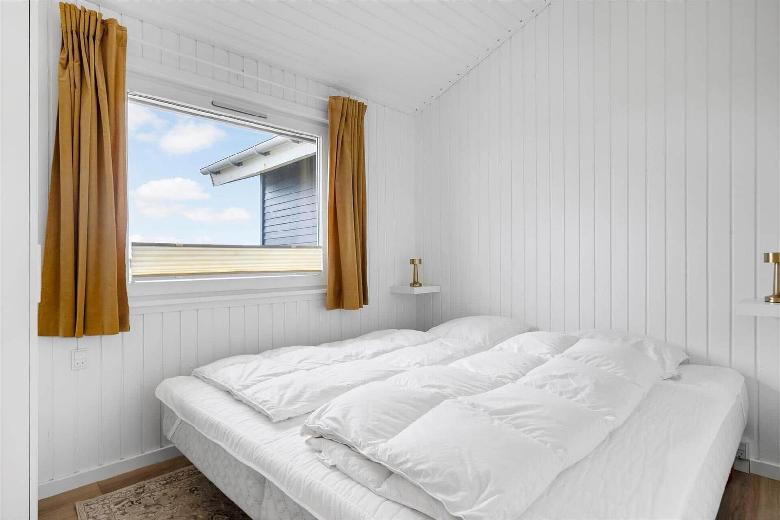 Schlafzimmer mit weißer Wandverkleidung, Bett mit weißen Bettwäsche und Fenster mit braunen Vorhängen.