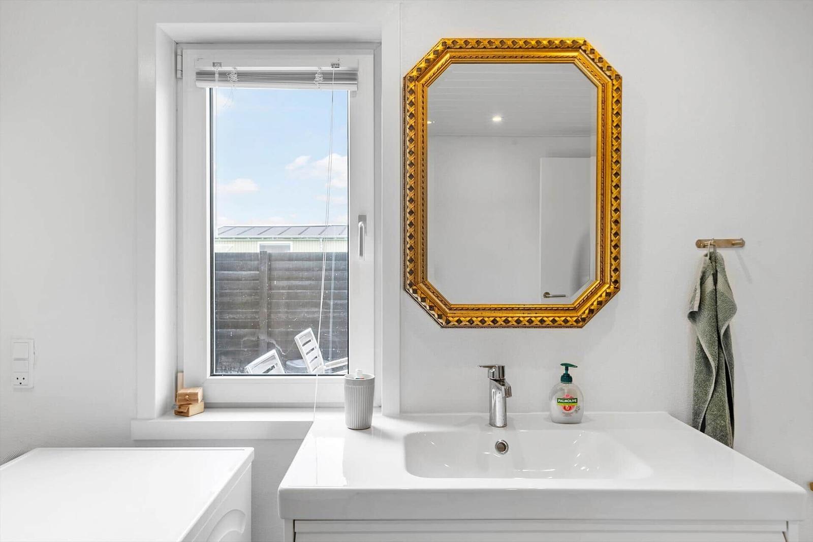 Badezimmer mit Waschbecken, goldgerahmtem Spiegel und Fenster zum Blick nach draußen.