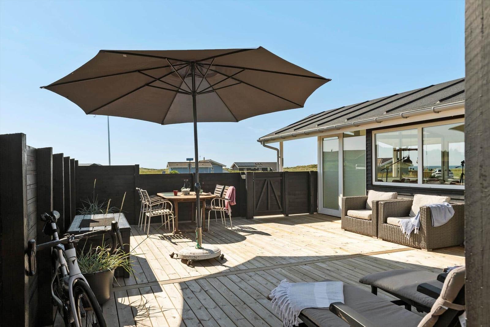 Terrasse mit Sonnenschirm, Tisch, Stühlen und Liegestühlen. Fahrrad und Pflanzen sind ebenfalls sichtbar.