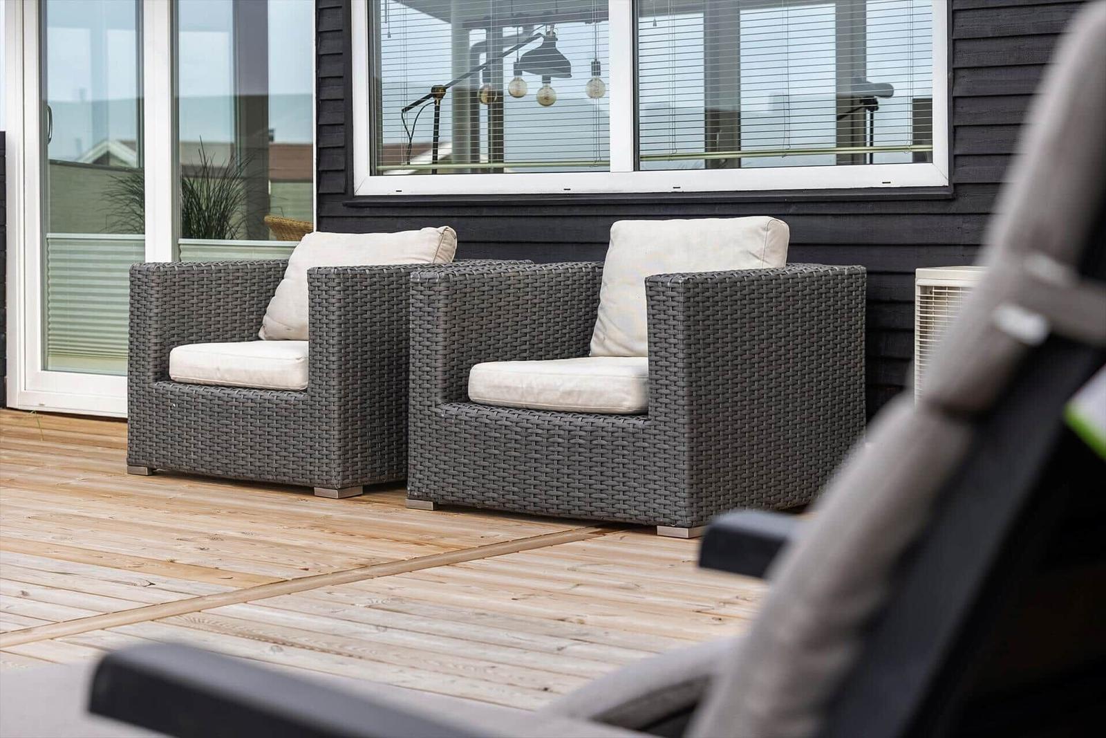 Zwei graue Rattan-Sessel mit weißen Kissen stehen auf einer Holzterrasse vor einem Haus.