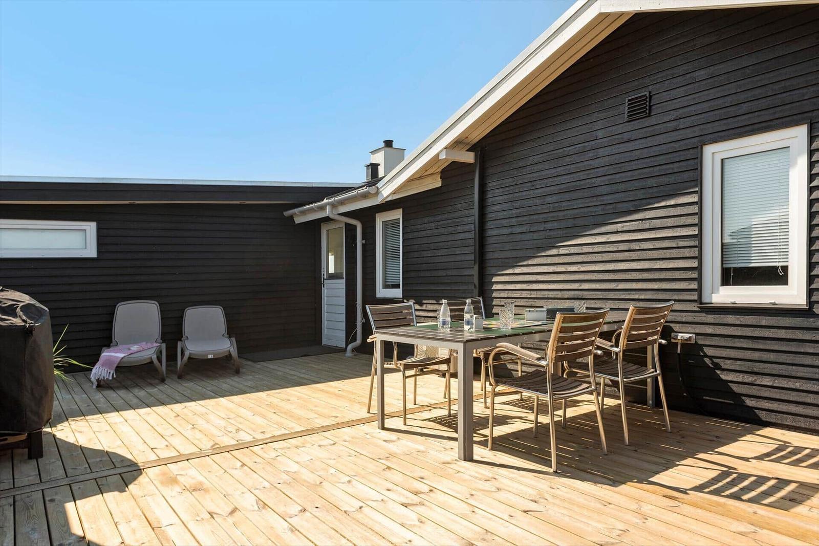 Terrasse mit Holzboden, Tisch, Stühlen und Liegestühlen an dunkelholzverkleidetem Haus.
