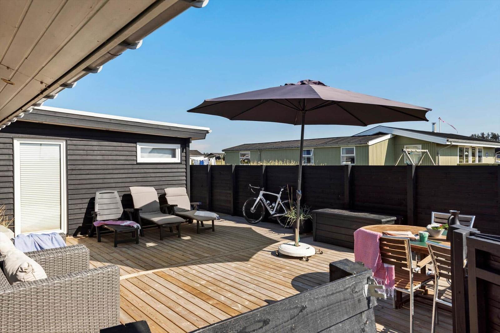 Terrasse mit Sonnenschirm, Liegestühlen und Tisch. Fahrrad und Holzgeländer sind sichtbar.