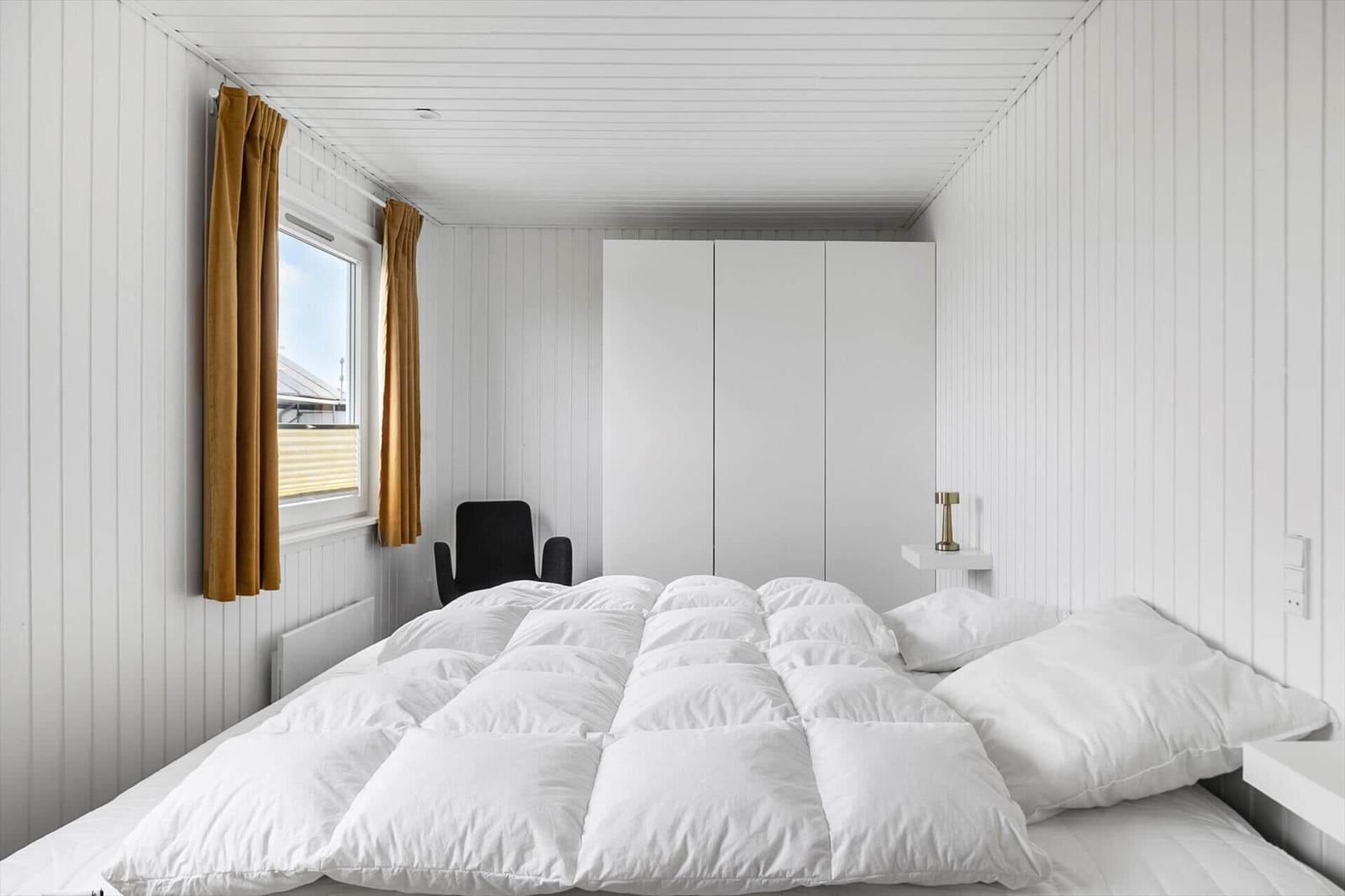 Schlafzimmer mit weißem Bett, Fenster mit braunen Vorhängen und weißen Wänden.